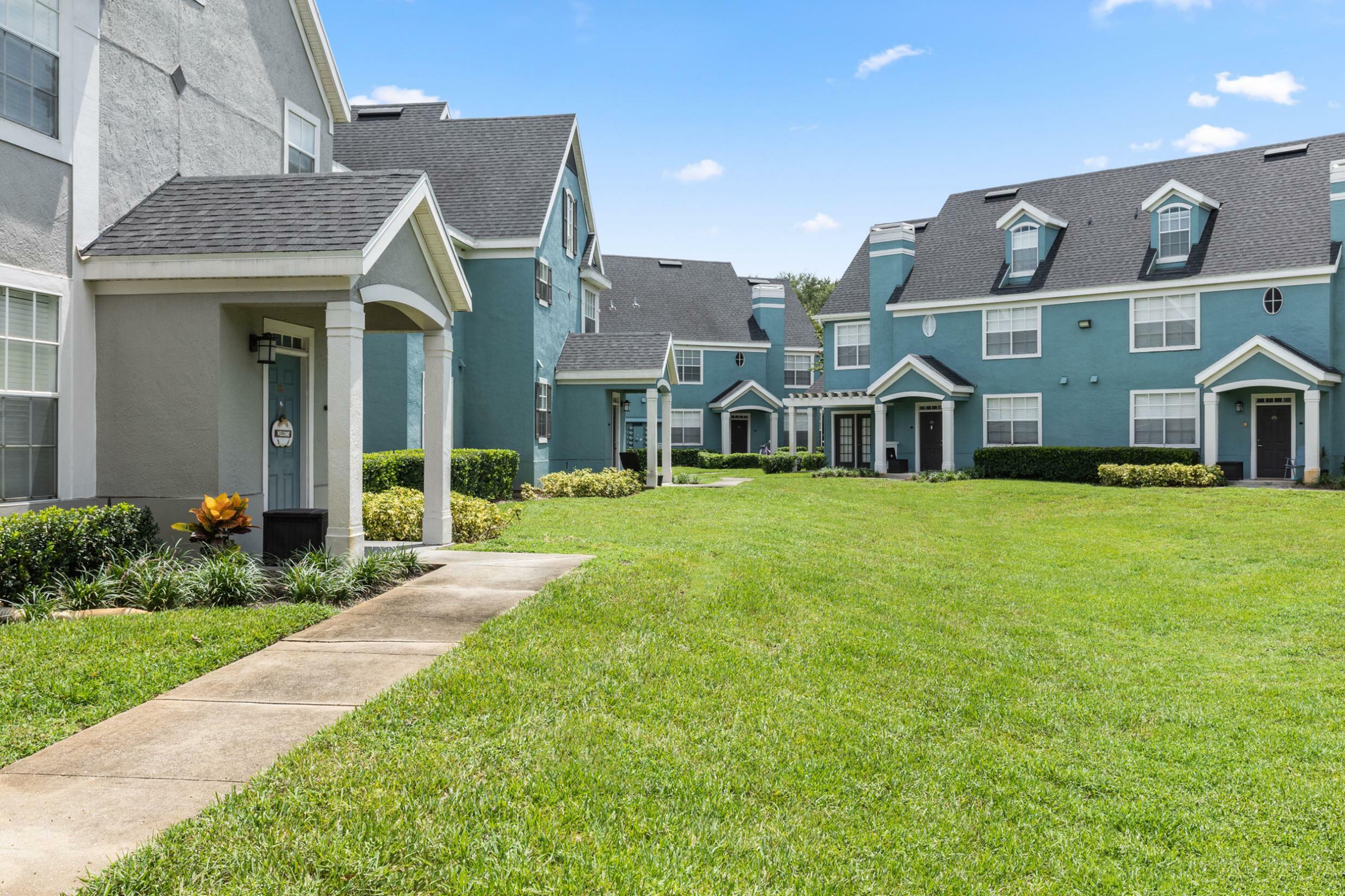 Charming townhouse community with well-manicured lawns, classic architectural details, and inviting blue and white facades under a clear blue sky.