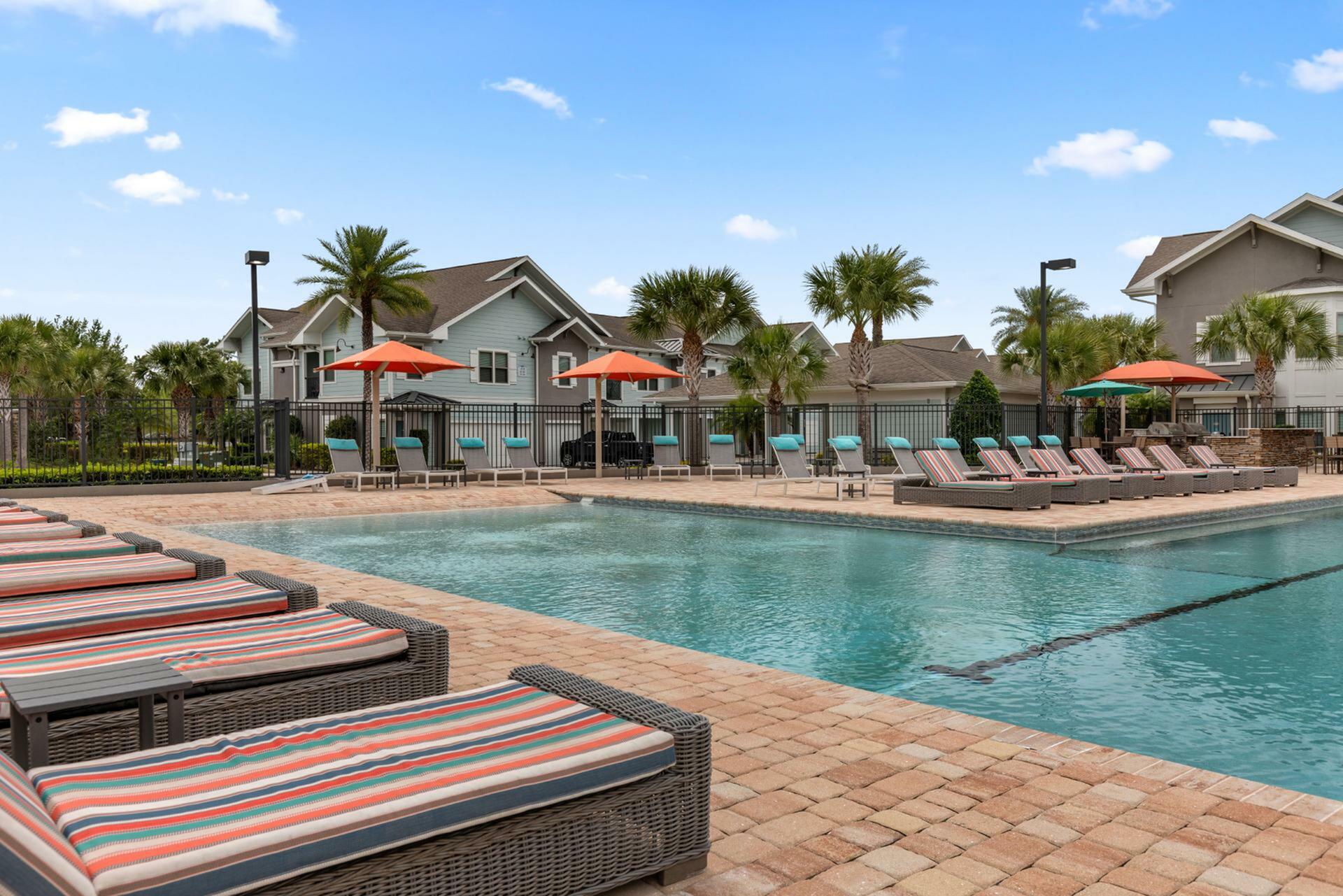 Luxurious poolside view at a modern apartment complex with relaxing sun loungers, colorful umbrellas, and vibrant palm trees under a bright blue sky.
