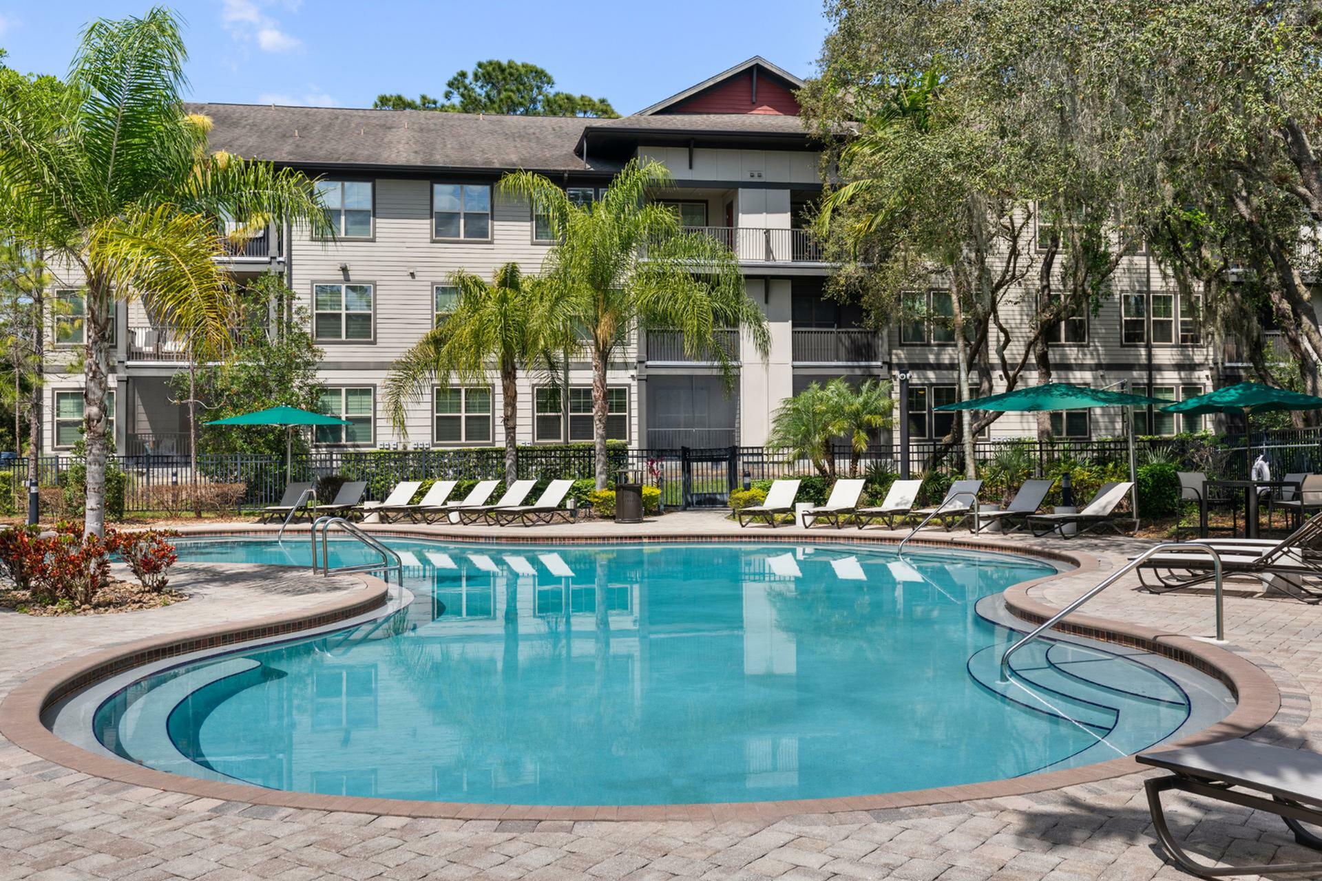 Charming apartment complex with a sunlit pool surrounded by lush palm trees and cozy lounge chairs, offering a serene and relaxing retreat.