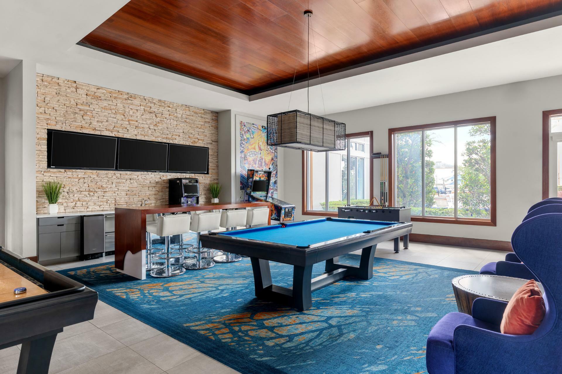 Luxurious apartment game room featuring a stylish pool table, modern bar area, and vibrant decor with large windows.