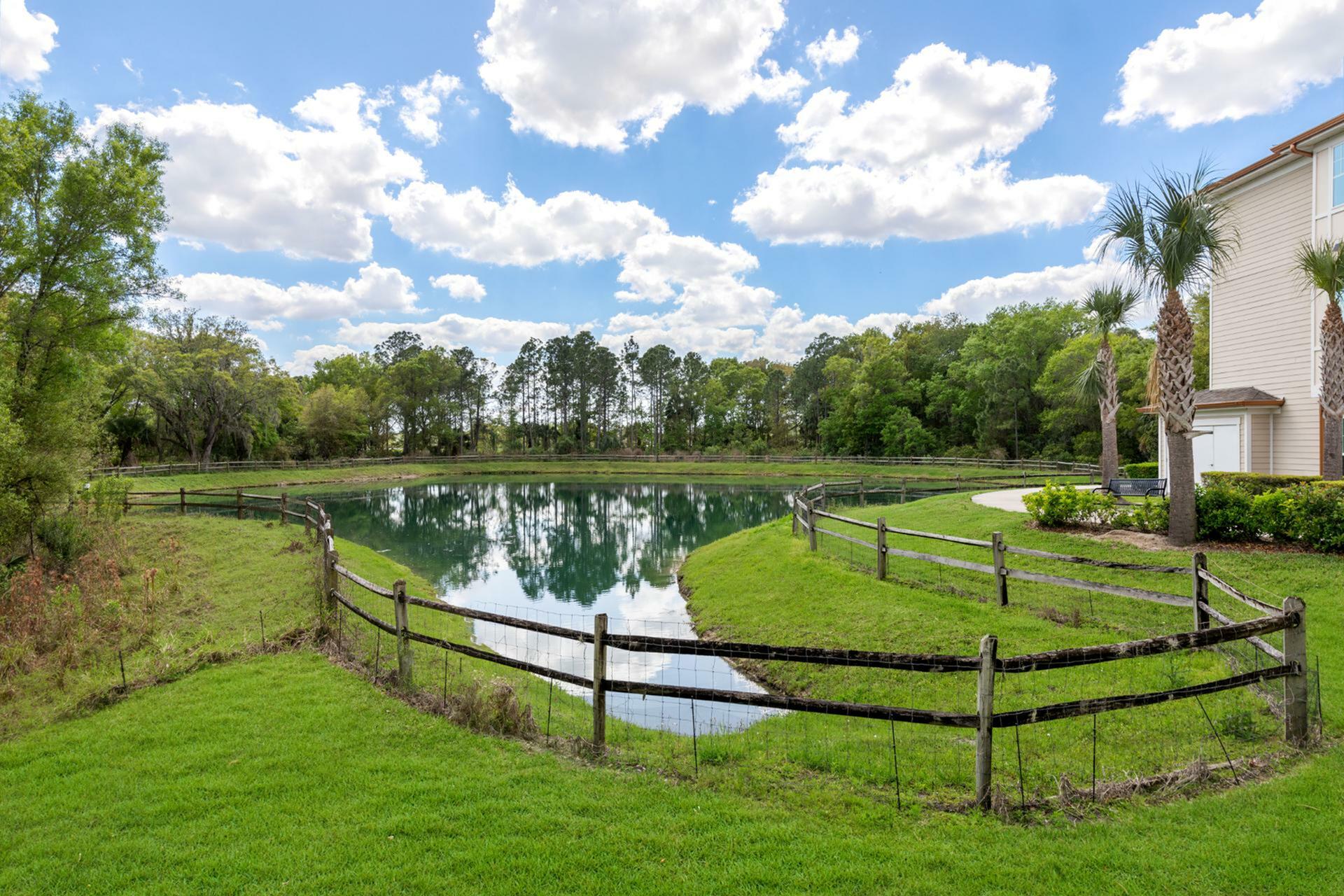 Tranquil lakeside view with lush green lawns, rustic wooden fence, and a charming residential building under a vibrant blue sky with fluffy clouds.
