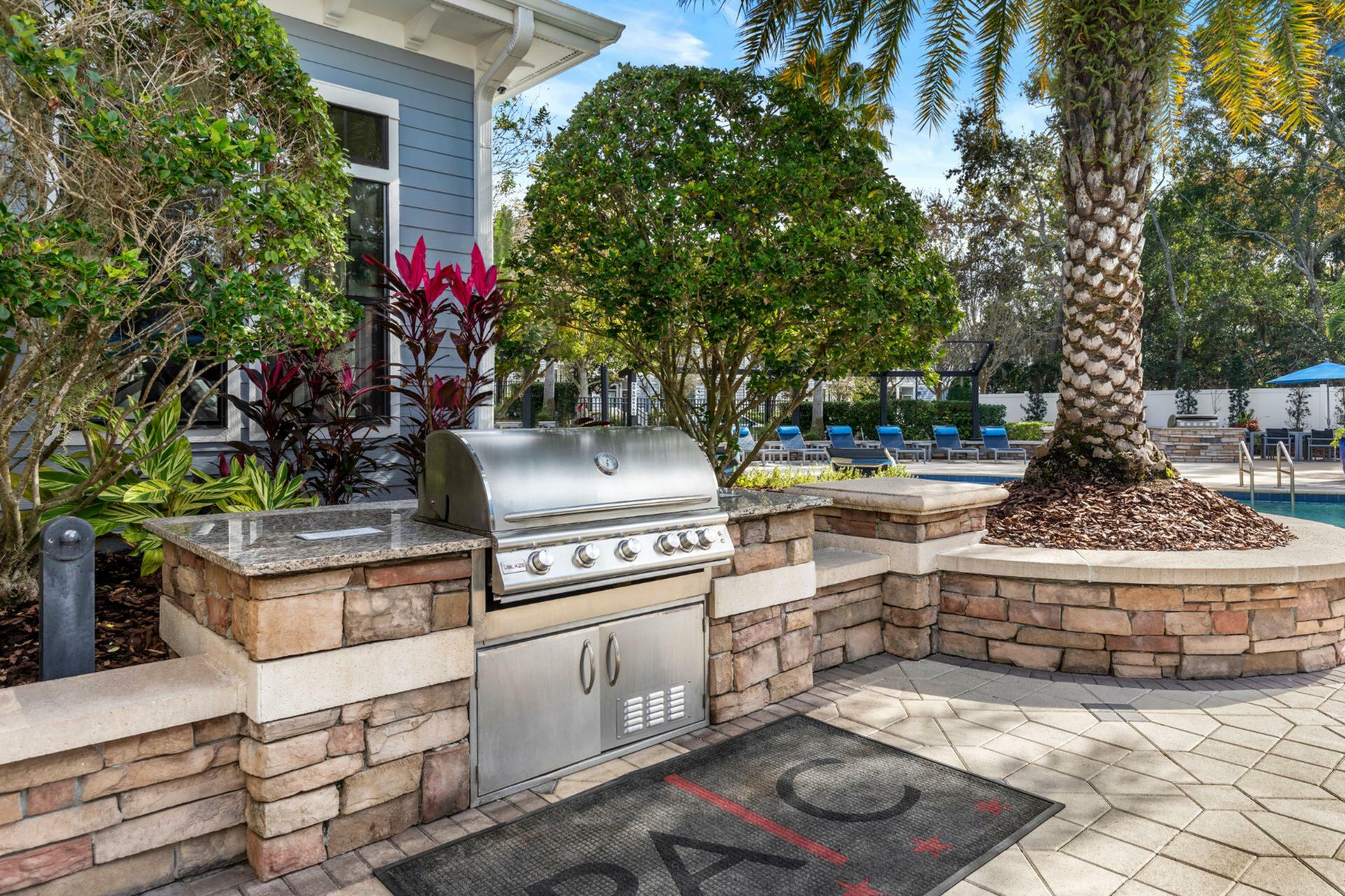 Inviting outdoor space with a stainless steel grill, lush greenery, and a sparkling pool nearby, perfect for entertaining.