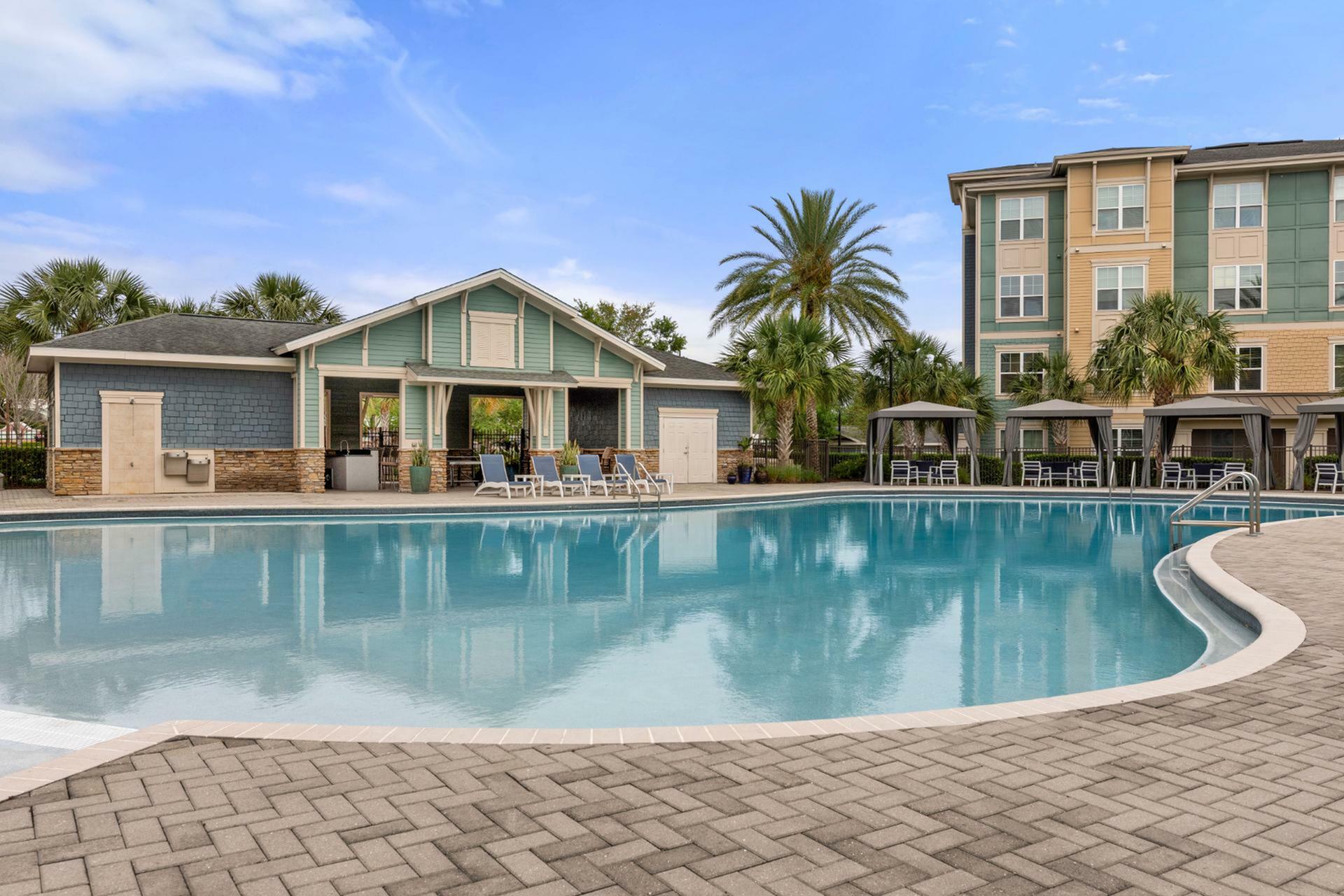Luxurious apartment setting featuring a sparkling outdoor pool, cozy cabanas, and lush palm trees under a clear blue sky.