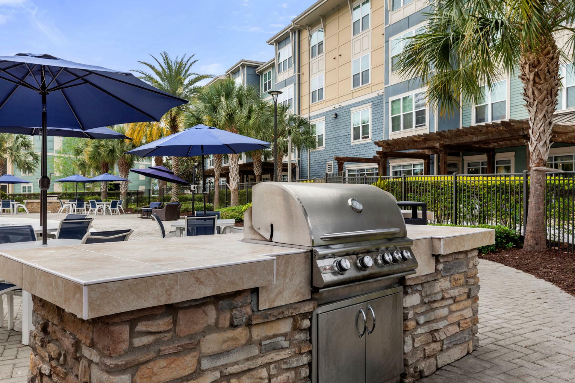 Chic outdoor patio area featuring a modern grill station, stylish seating under blue umbrellas, and lush greenery.