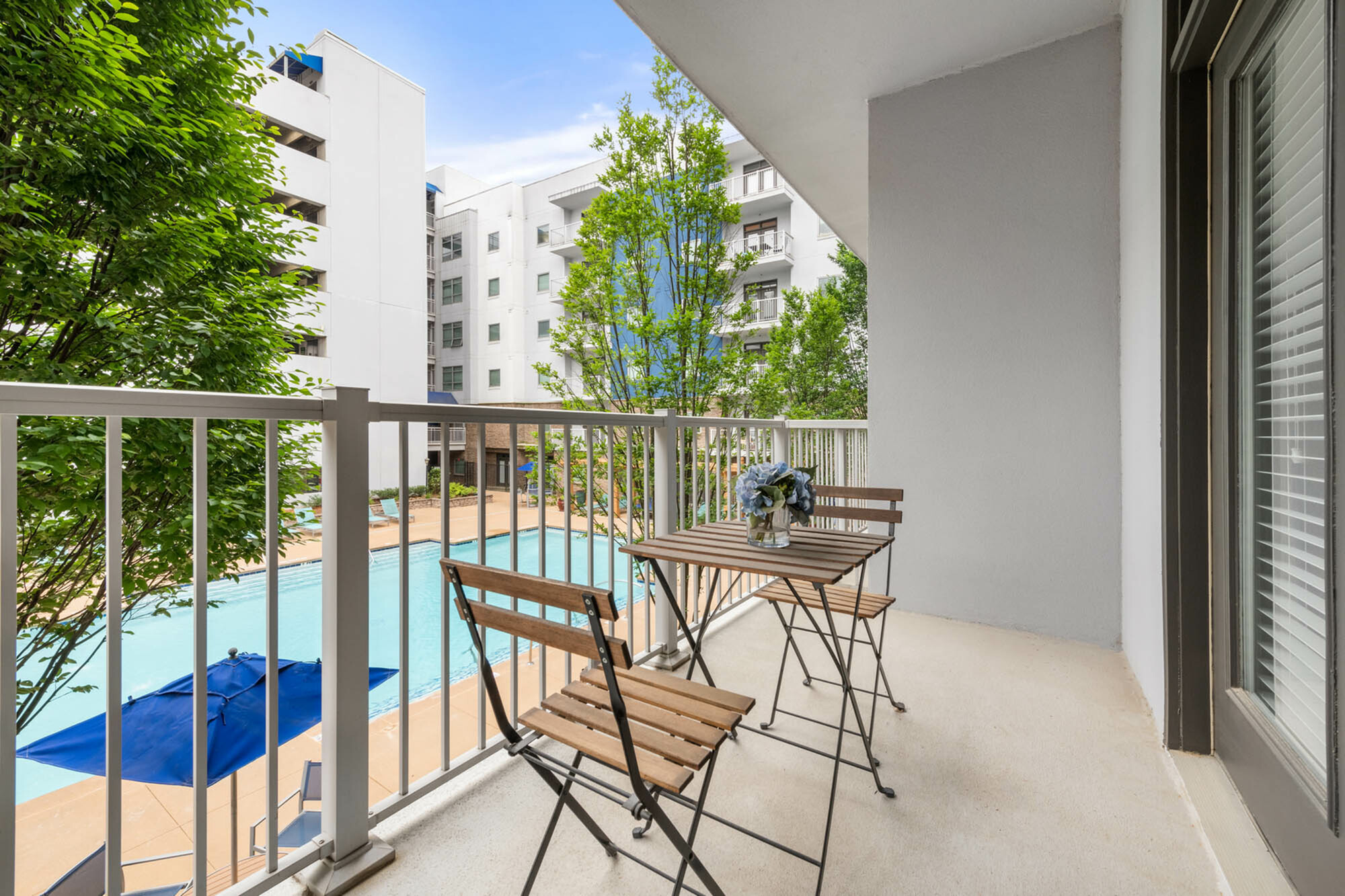 Inviting balcony view overlooking a pristine swimming pool, surrounded by lush greenery and modern architecture, complete with a charming outdoor bistro set.
