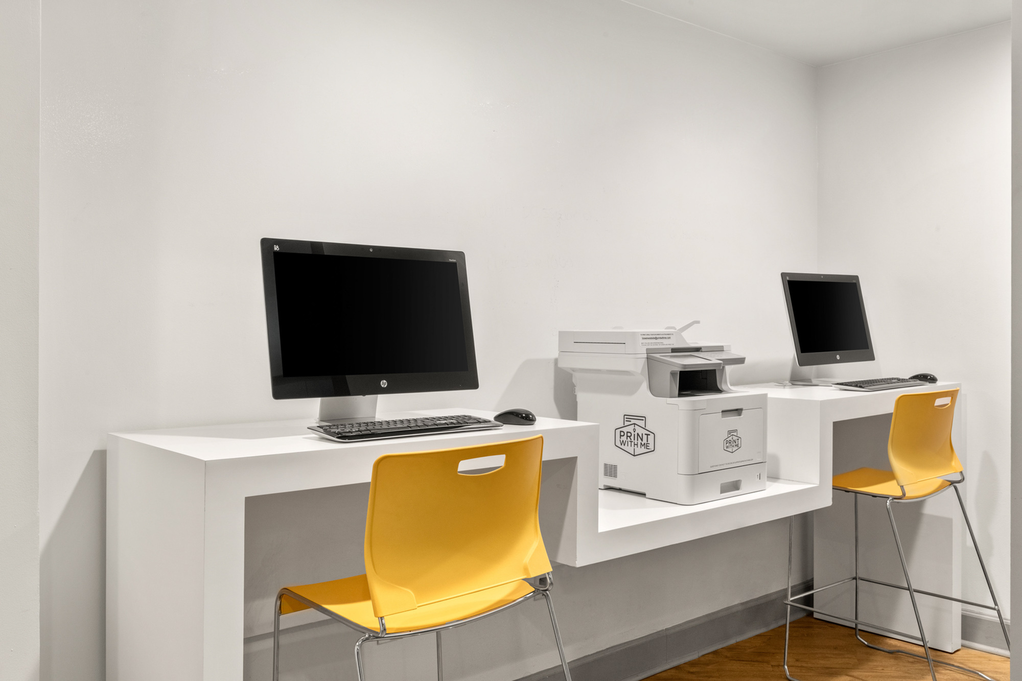 Modern shared office space with two sleek computers, a printer, and vibrant yellow chairs against a clean white backdrop.