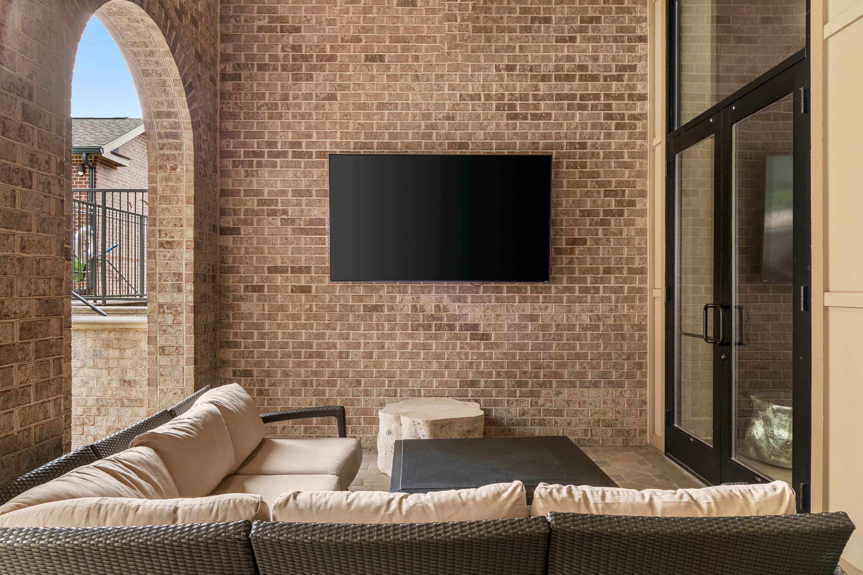 Chic outdoor patio with cozy wicker seating, modern TV, and elegant brick archway.