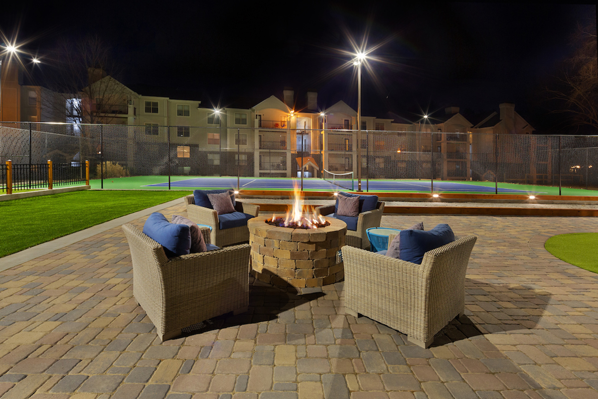 Elegant outdoor lounge area with cozy wicker chairs around a glowing fire pit, overlooking a well-lit tennis court, perfect for nighttime gatherings.