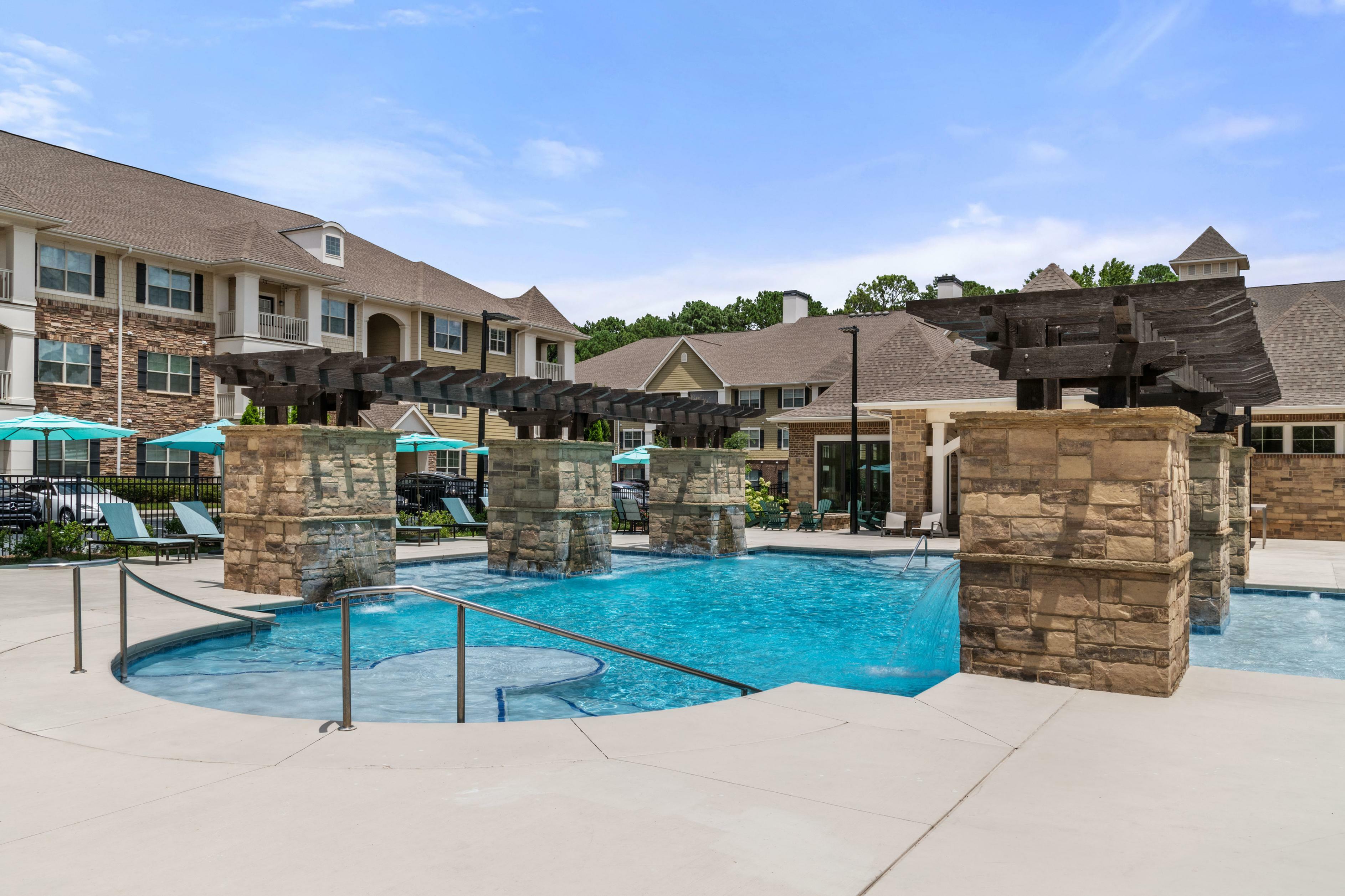 Luxurious apartment complex with a stunning pool area, featuring stone pillars and lounge chairs under blue umbrellas, perfect for relaxation.