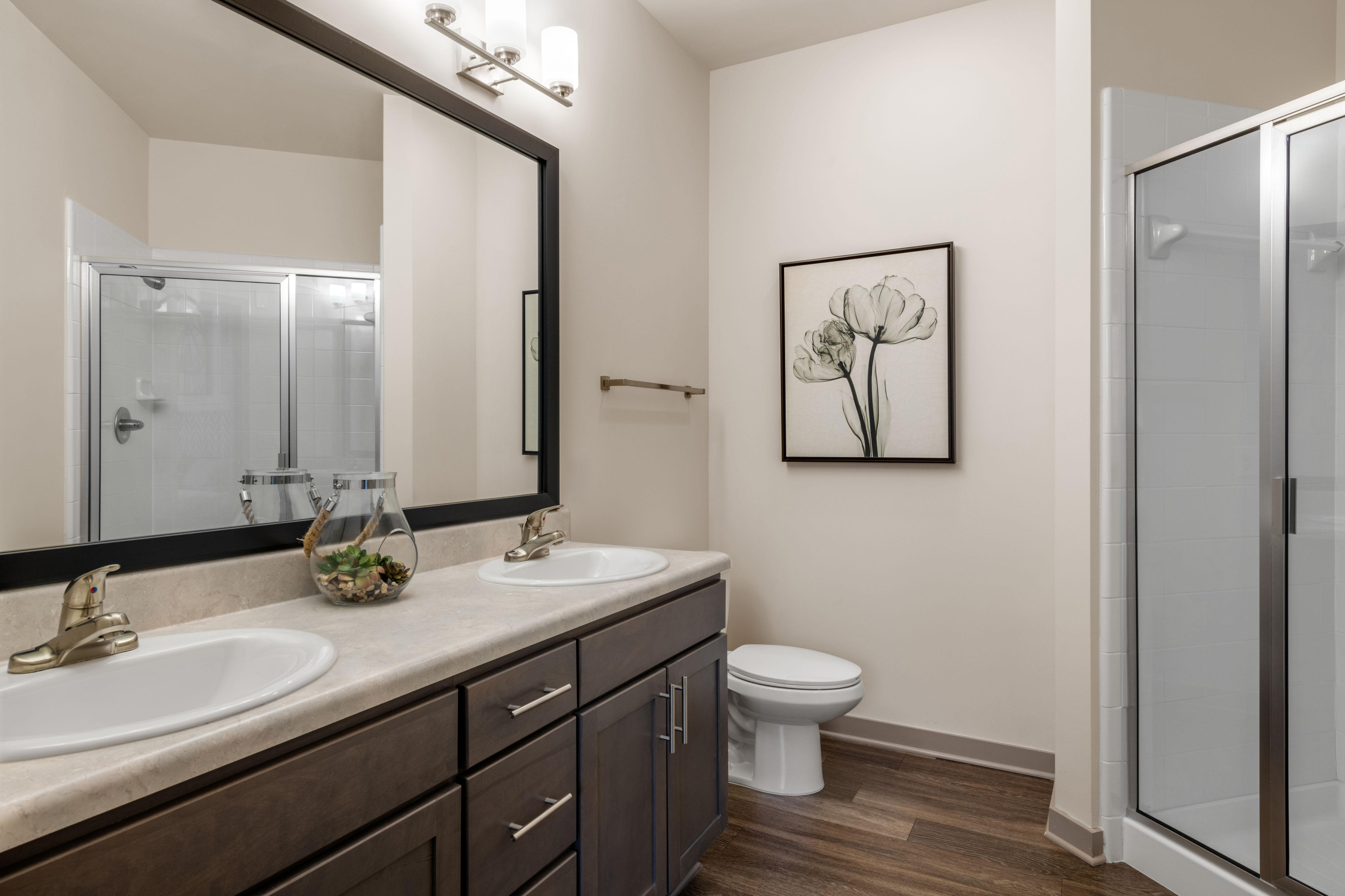Elegant bathroom design featuring dual sinks with modern fixtures, stylish dark wood cabinetry, framed floral artwork, and a spacious glass-enclosed shower.