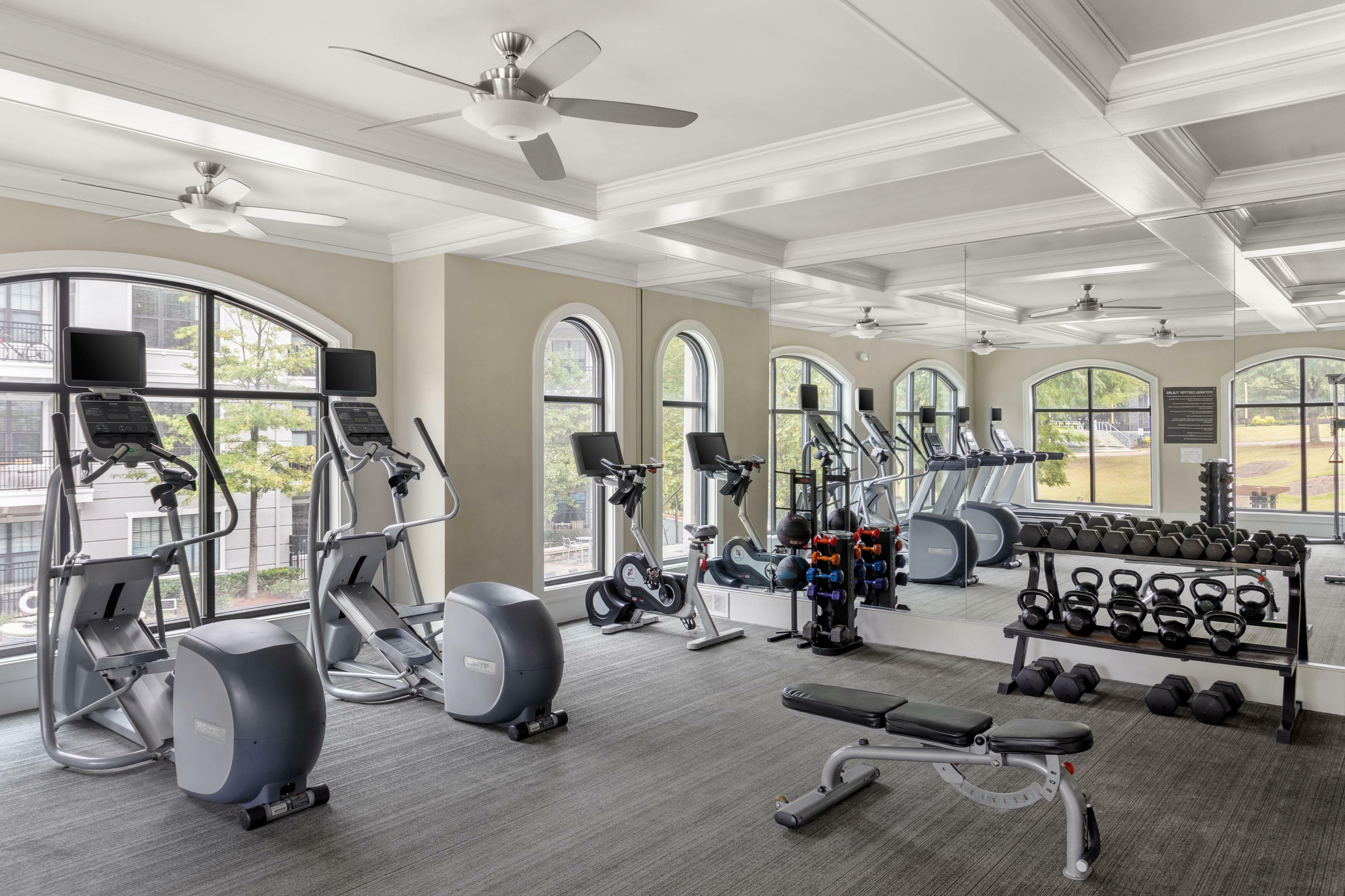 Modern fitness center with state-of-the-art equipment, featuring elliptical machines, free weights, and a mirrored wall under elegant ceiling fans and large arched windows.