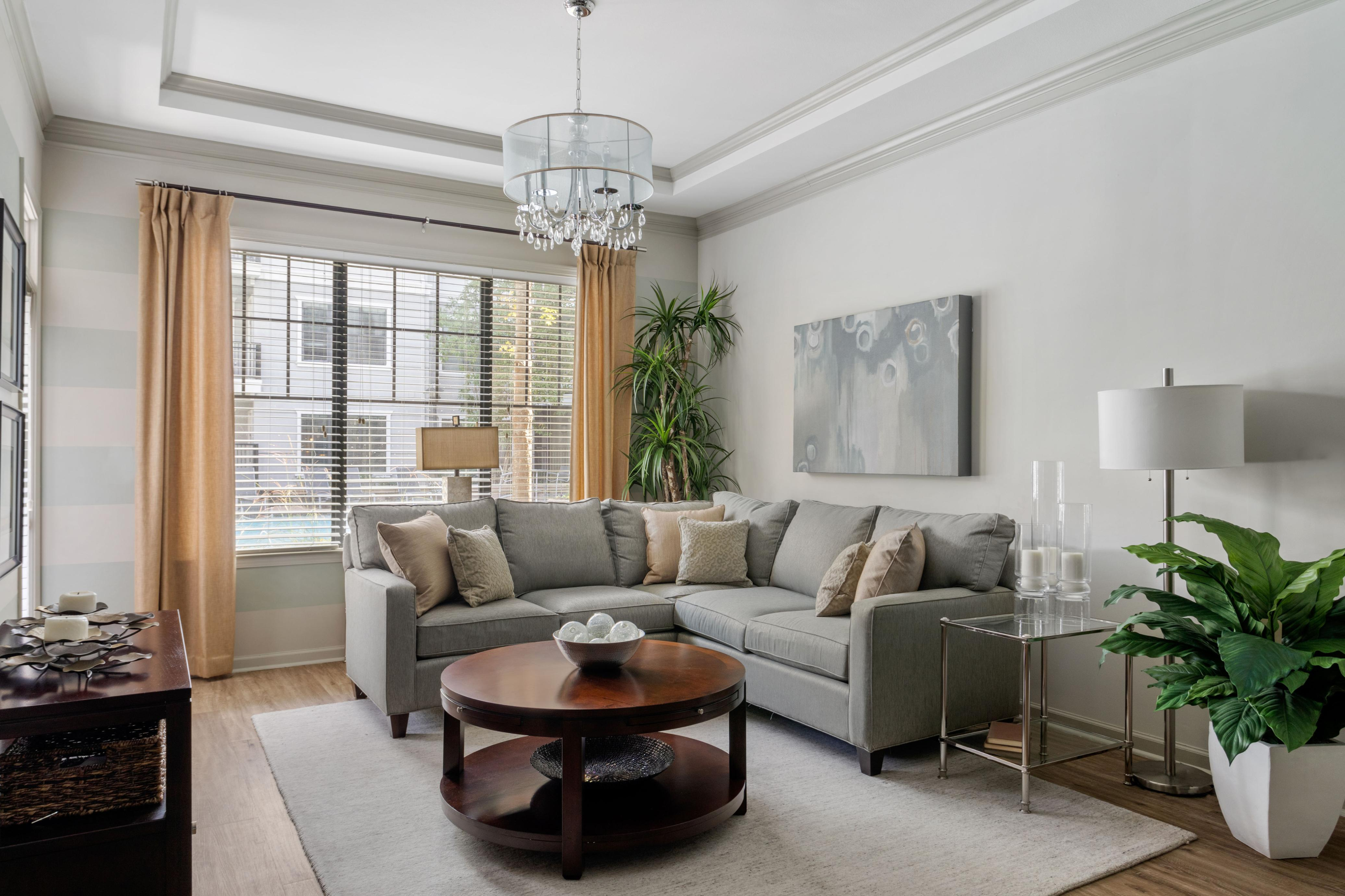 Inviting living room with elegant gray sectional sofa, stylish chandelier, and abundant natural light from large windows, complemented by modern decor and lush greenery.