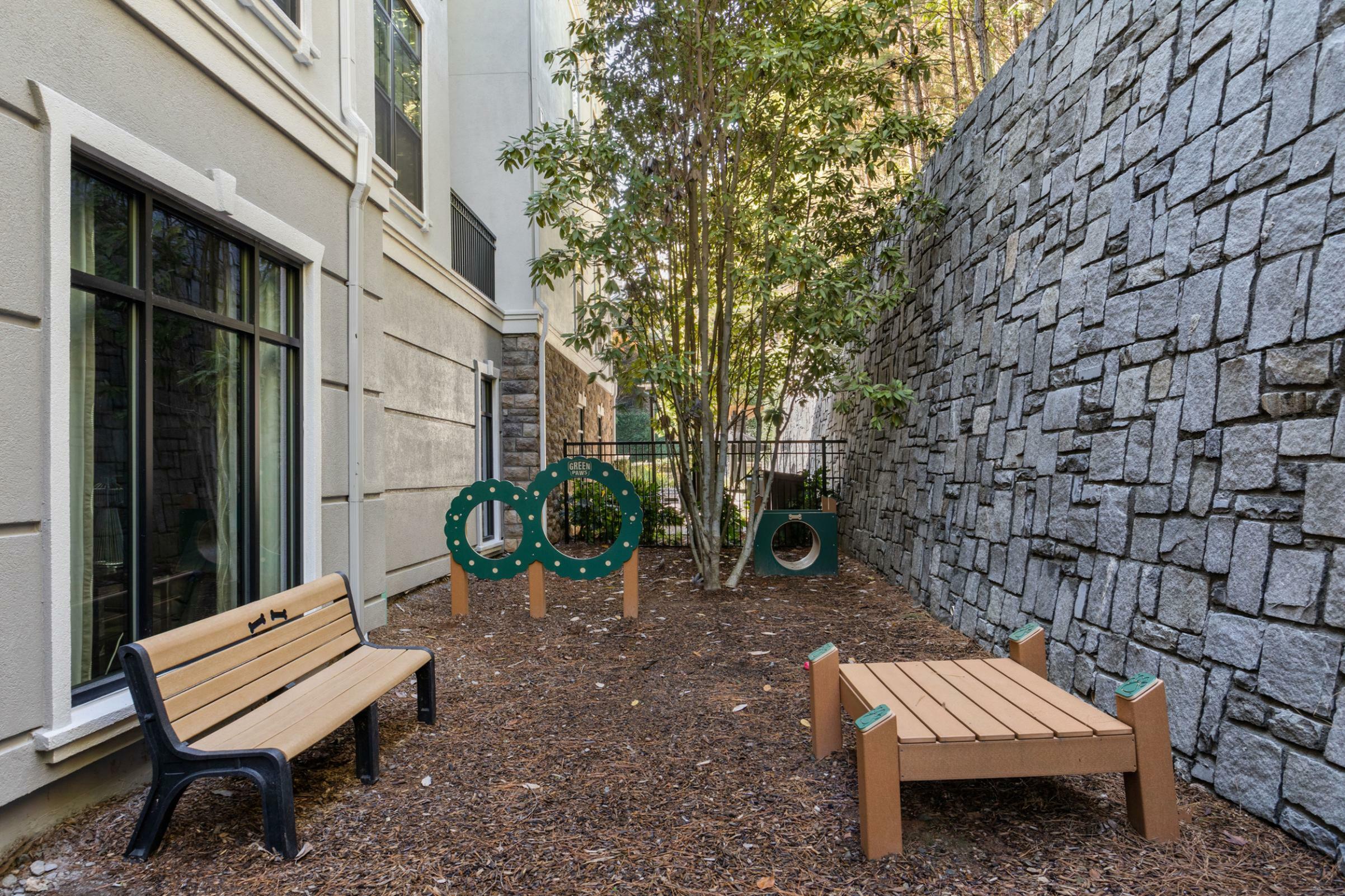 Charming outdoor space featuring a cozy bench, modern stone wall, and playful pet agility equipment surrounded by lush greenery.