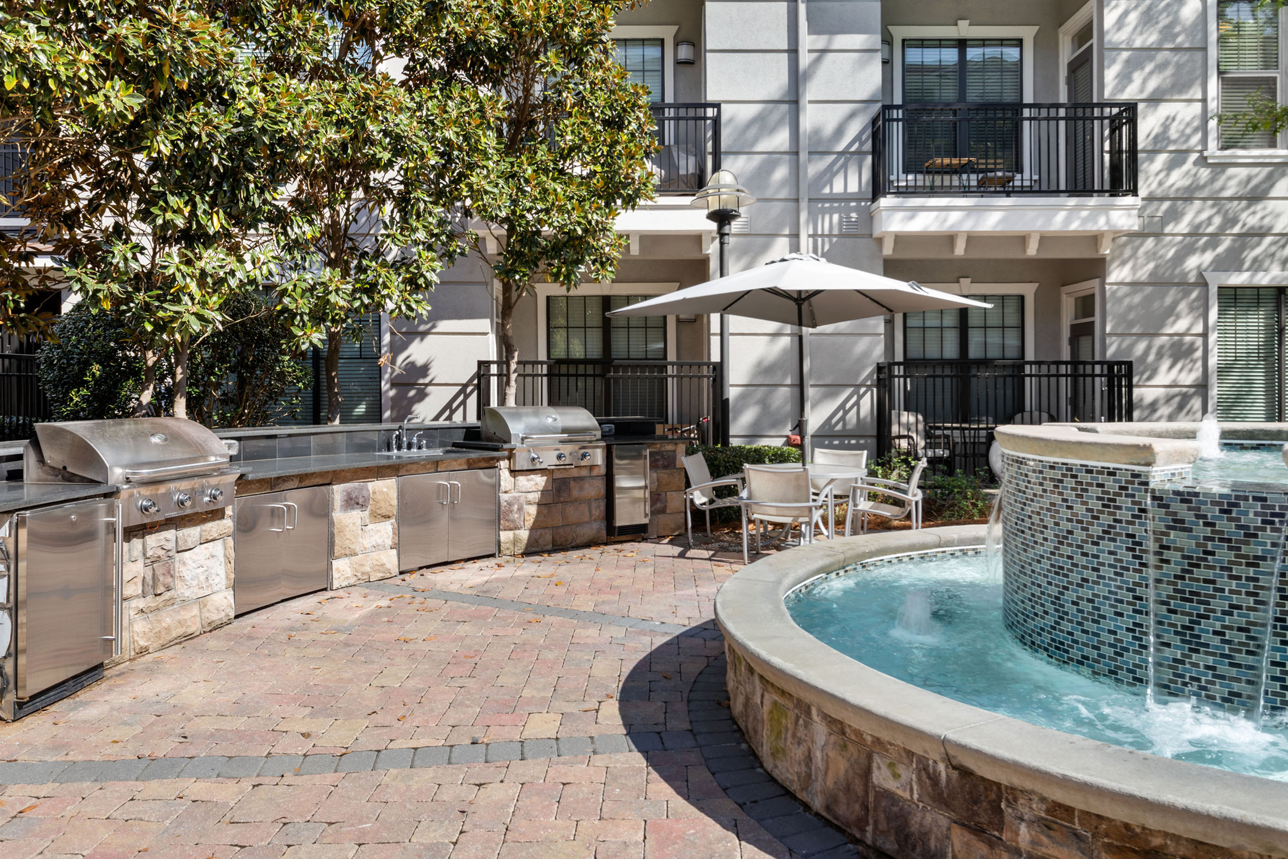 Luxurious outdoor courtyard with modern barbecue grills, elegant seating under umbrellas, and a stunning mosaic water fountain.
