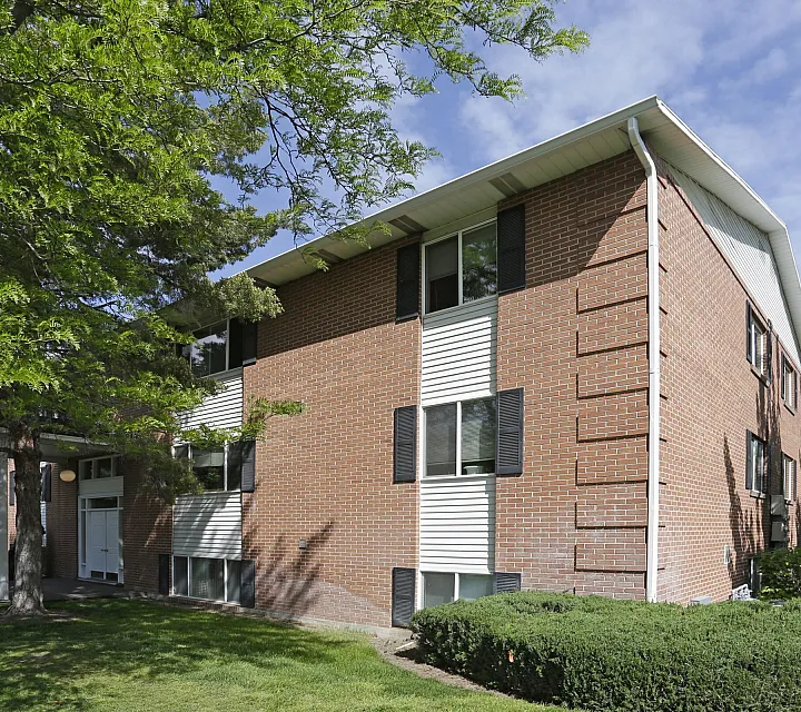 Corner of a brick building with lush trees and hedges at Linden Colonial in Holladay, UT