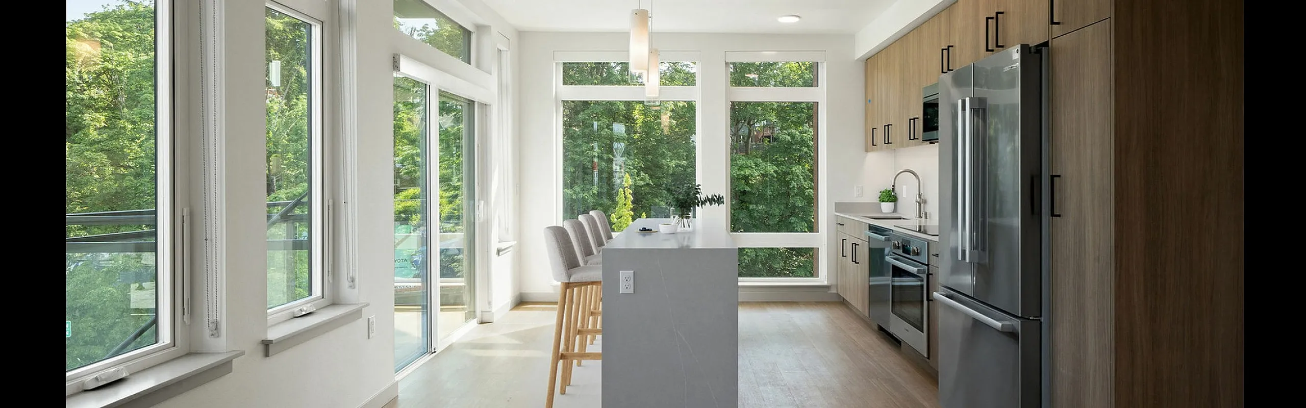 Bright modern kitchen with sleek stainless steel appliances, large windows, and an elegant island with high chairs, surrounded by lush greenery views.