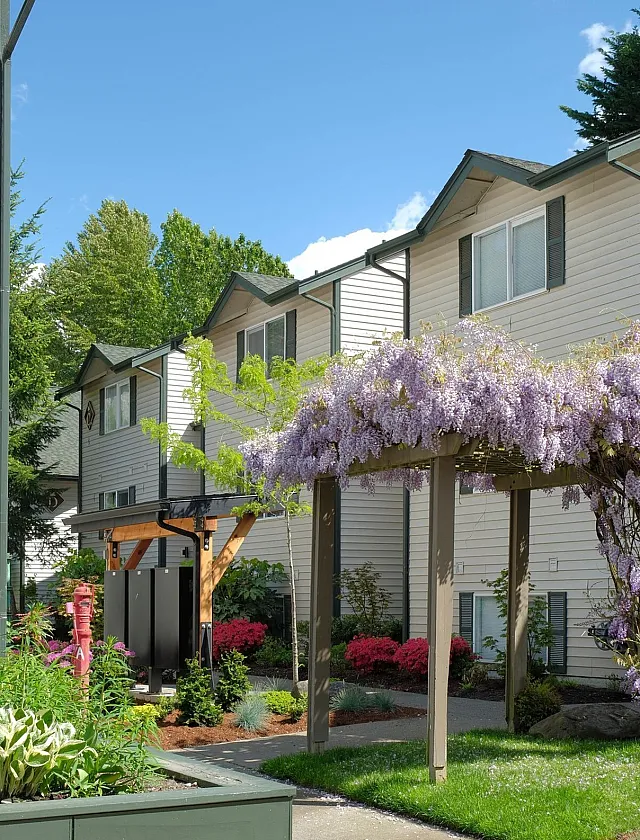 Charming apartment exterior with lush landscaping, vibrant flowers, and a picturesque pergola adorned with purple wisteria.