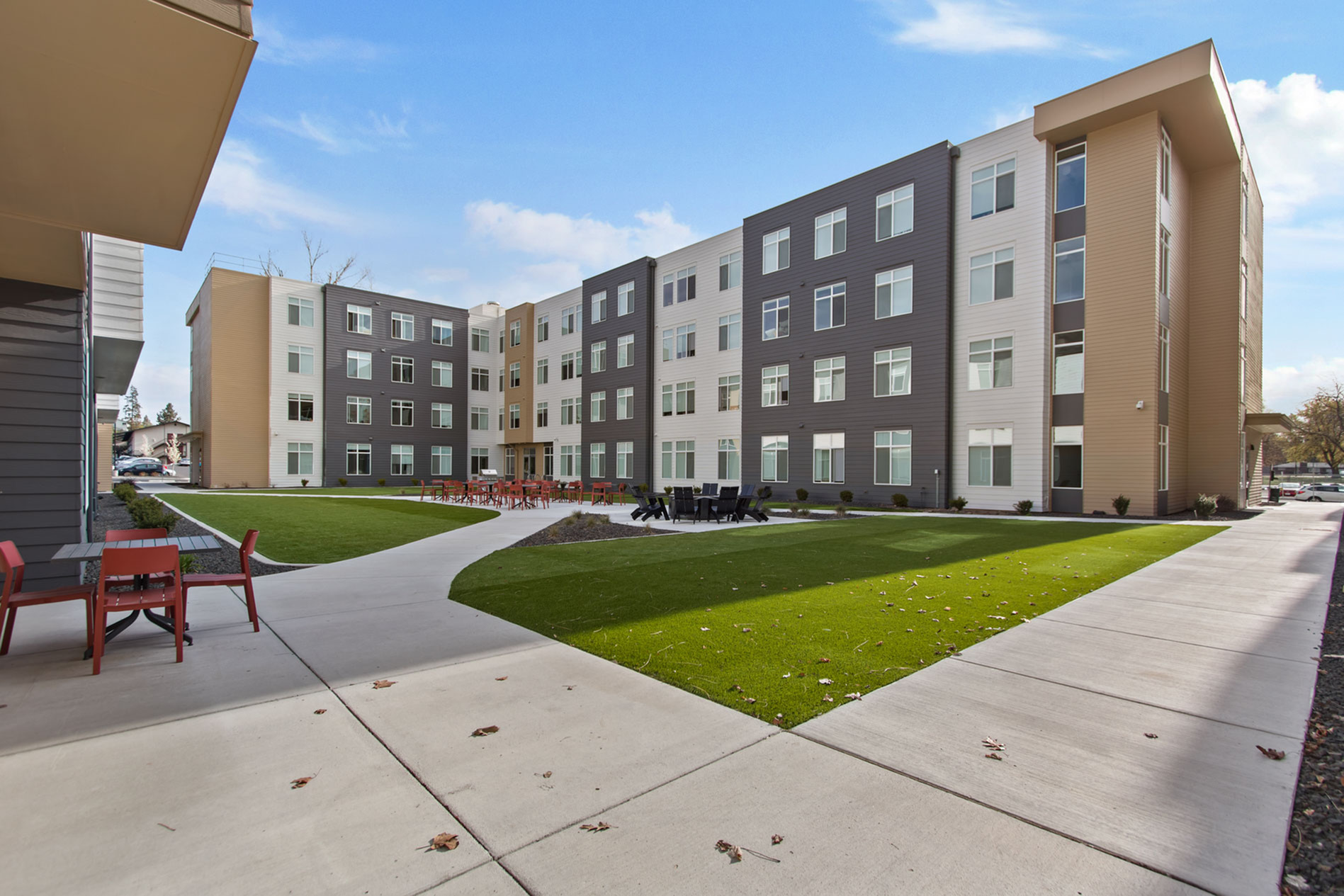 Modern apartment complex with stylish architecture, featuring a spacious courtyard, lush green lawns, and inviting outdoor seating area under a clear blue sky.