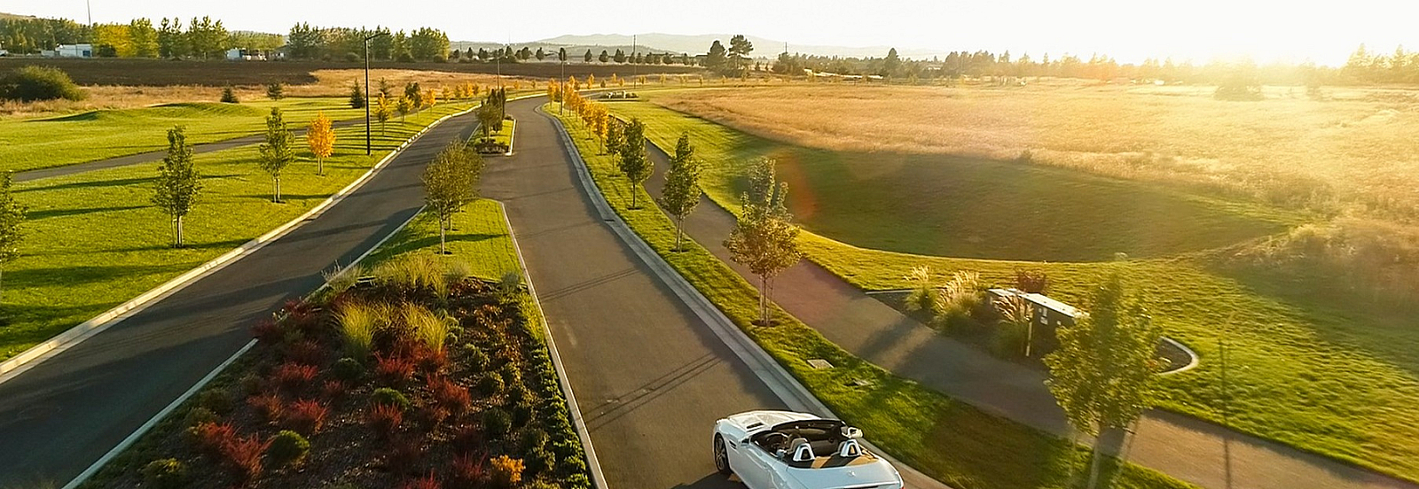 Serene suburban road lined with lush greenery and a convertible cruising at sunset, offering a picturesque and peaceful setting.