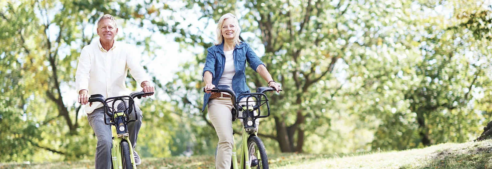 Joyful couple riding bicycles on a sunny park path, surrounded by lush green trees and vibrant nature.