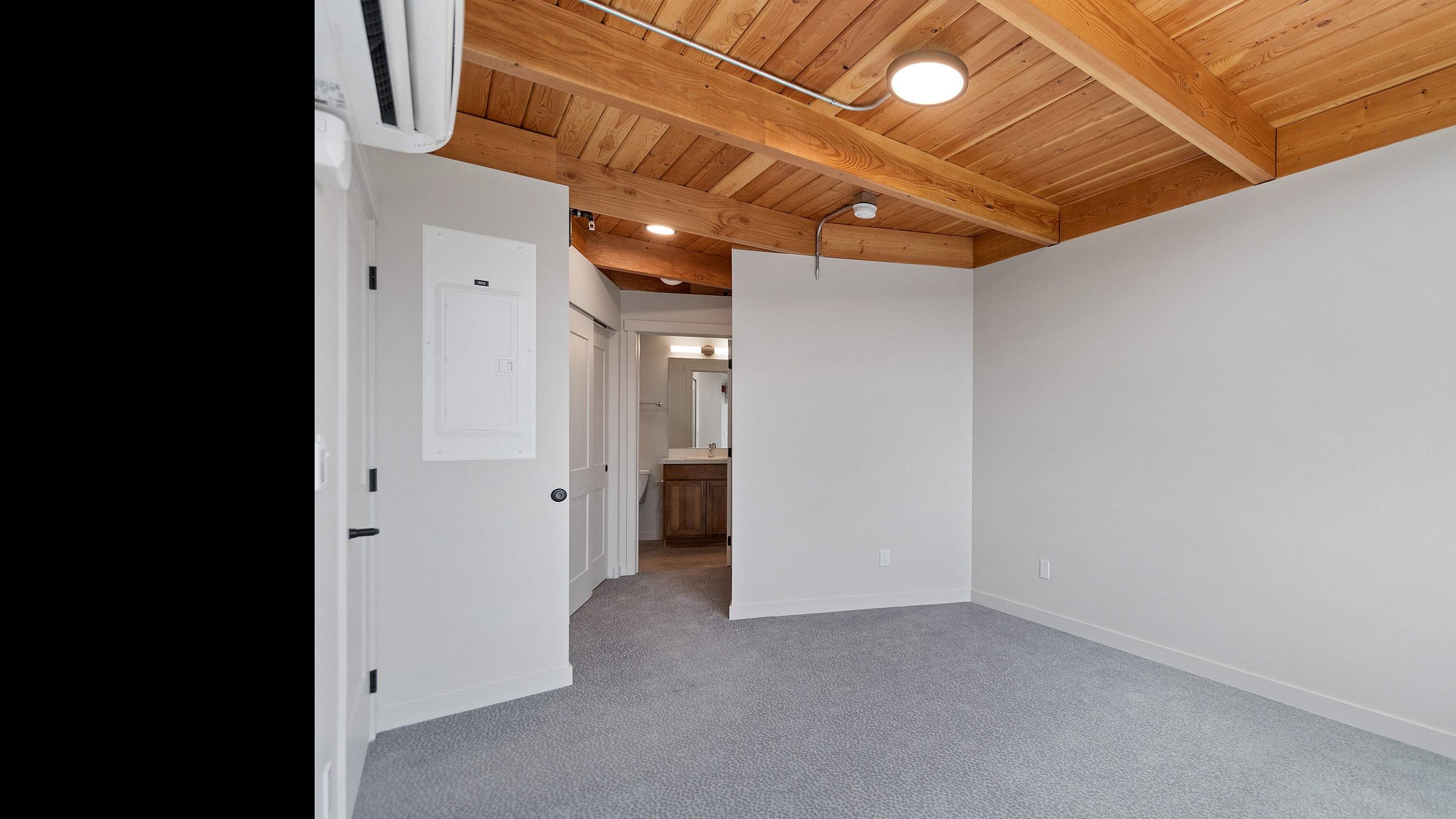 Bright, modern apartment with elegant wood beam ceilings, cozy carpeted flooring, and a sleek, inviting bathroom entrance.