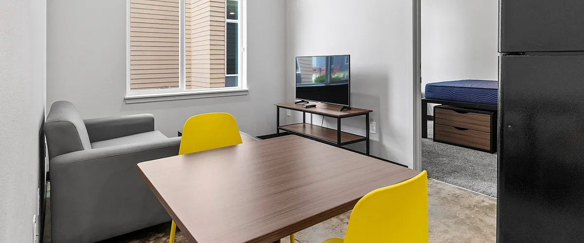 Modern apartment living area with sleek design, featuring a comfy gray sofa, vibrant yellow chairs, a polished wooden table, and a flat-screen TV, leading into a cozy bedroom.