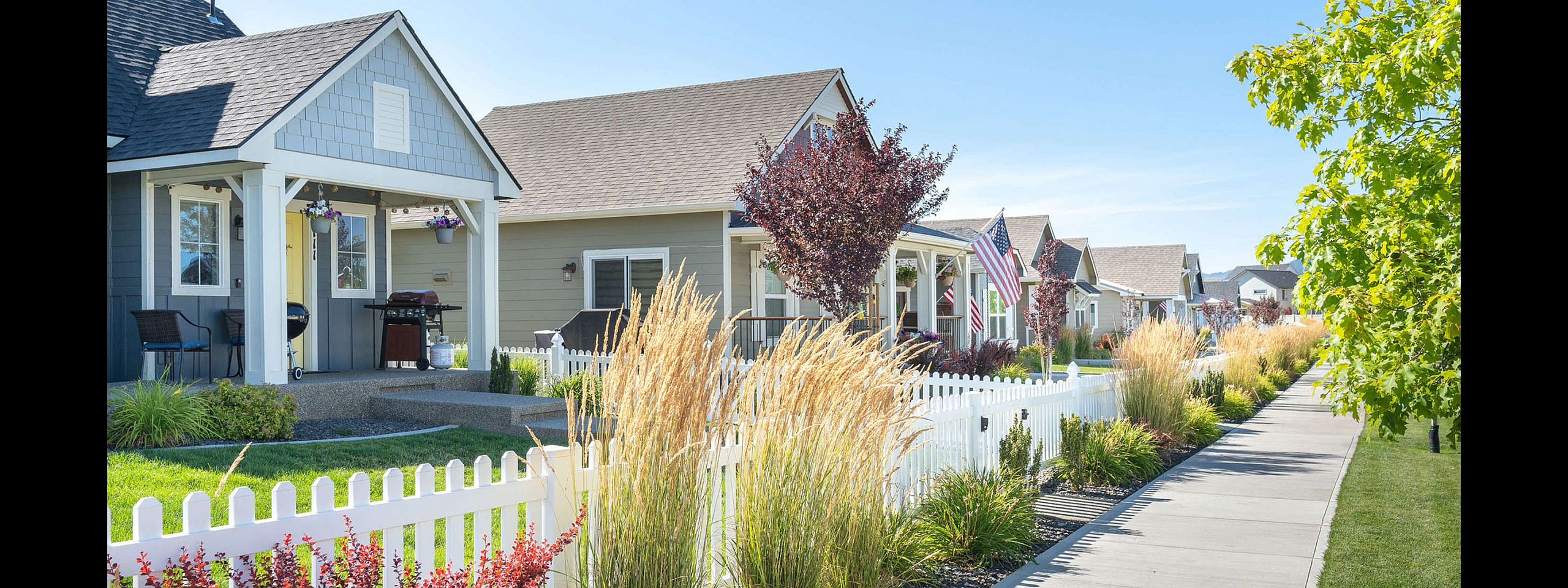 Charming suburban neighborhood with cozy houses, white picket fences, and lush landscaping under a bright blue sky.