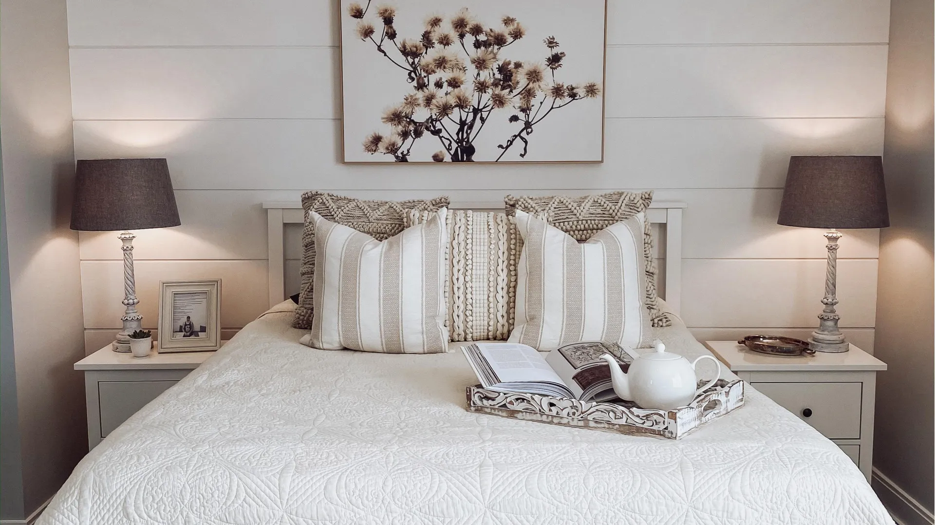 Queen bed layered with linens, two bedside tables and lamps to the side and a colorful piece of art hanging on the wall above the bed.
