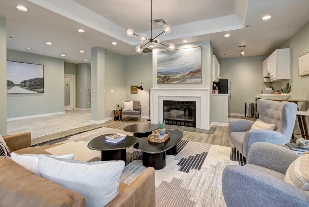 Clubhouse interior photo, showing white fireplace with large beach painting above. Light brown leather sofa faces the fire place, with two gray wingback chairs beside and large, modern coffee table in the center. Kitchenette in the back is