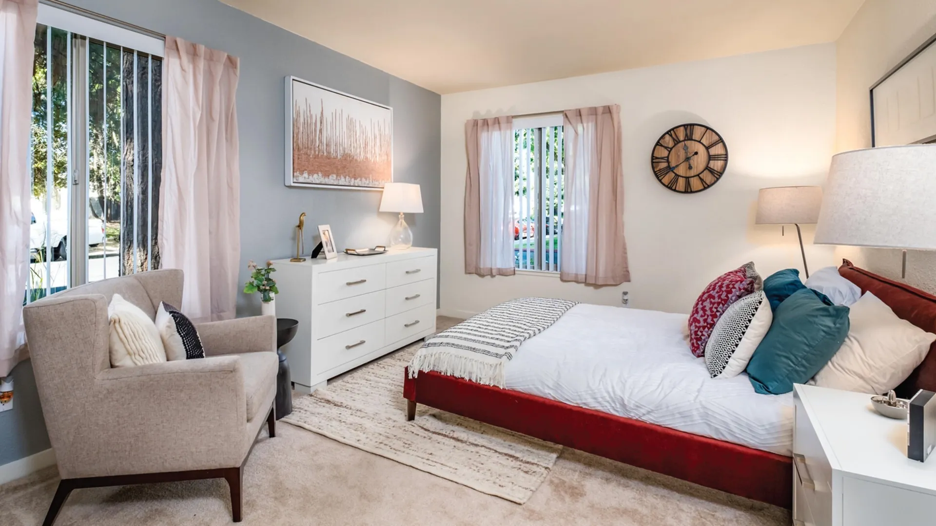 Bedroom with light carpeting, queen sized bed with two end tables, dressers, two windows, and living chair.