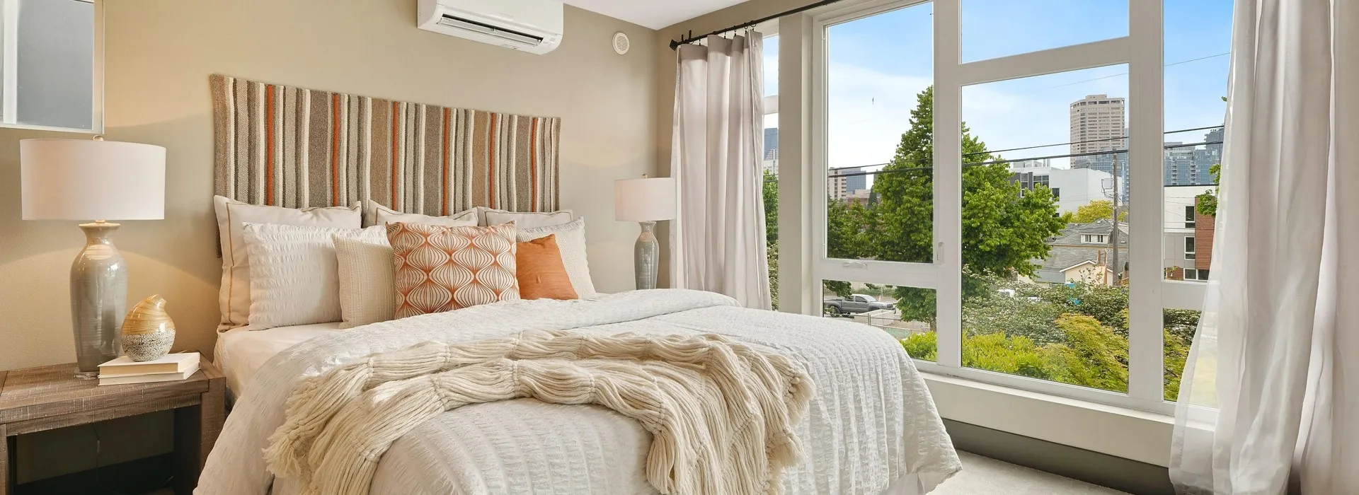 Warm toned room with white bedsheets and orange accent pillows viewing into neighborhood through floor to ceiling windows