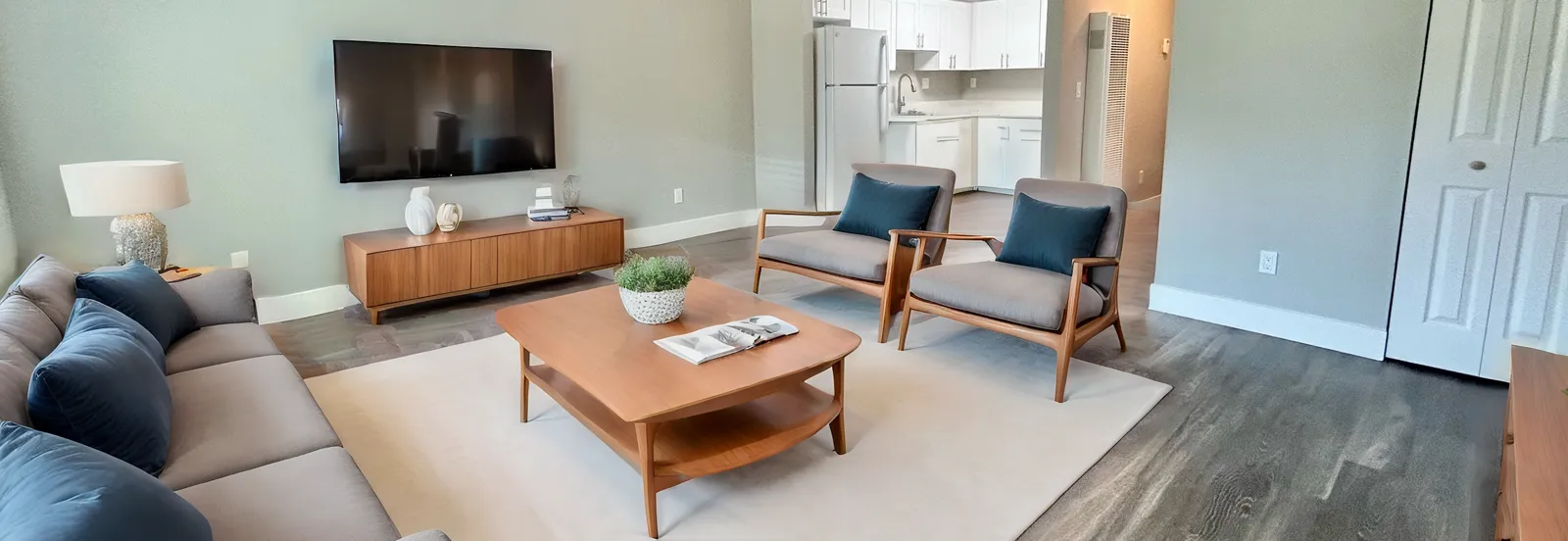 Furnished living room with TV, brown coffee table, two light brown chairs and blue throw pillows.