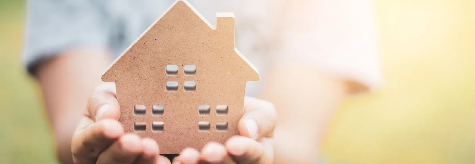 hands holding a wooden house figure in beaming light in the background creating a warm and inviting atmosphere