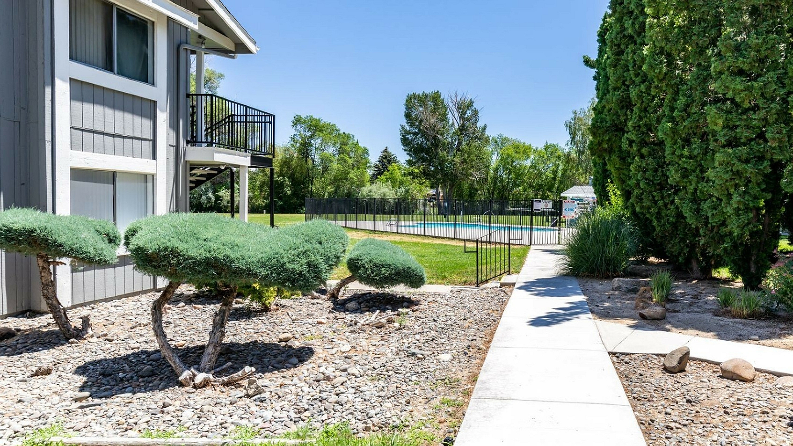 pathway with landscaping on each side leading up to black fence surrounding pool. blue skies create a vibrate atmosphere where the sun creates shadows of trees on pathway