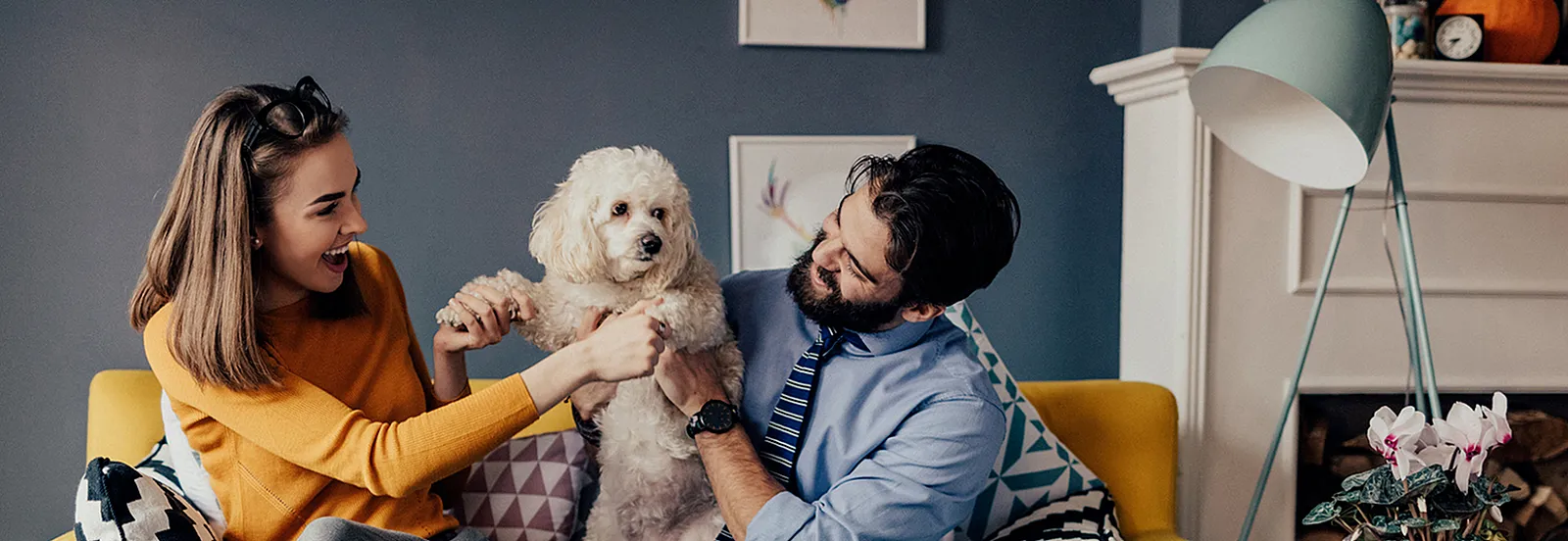 A man and woman sitting on a yellow couch holding up a white dog