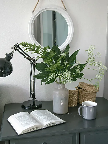A lamp, vase with faux flowers, and an open book on top of a grey dresser