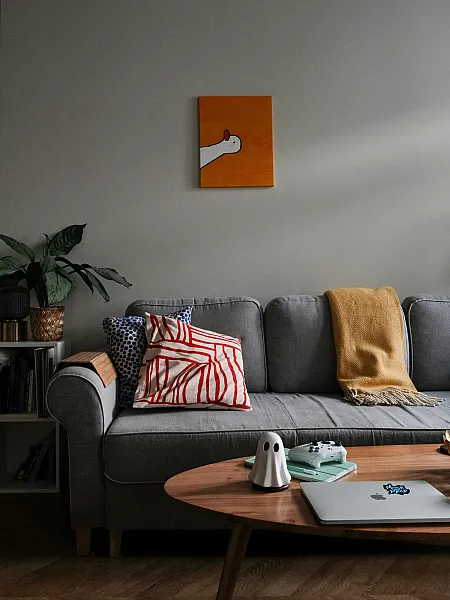 Cozy living room with a gray sofa adorned with patterned pillows and a mustard throw. A quirky goose painting hangs above, and a coffee table holds books and decor.