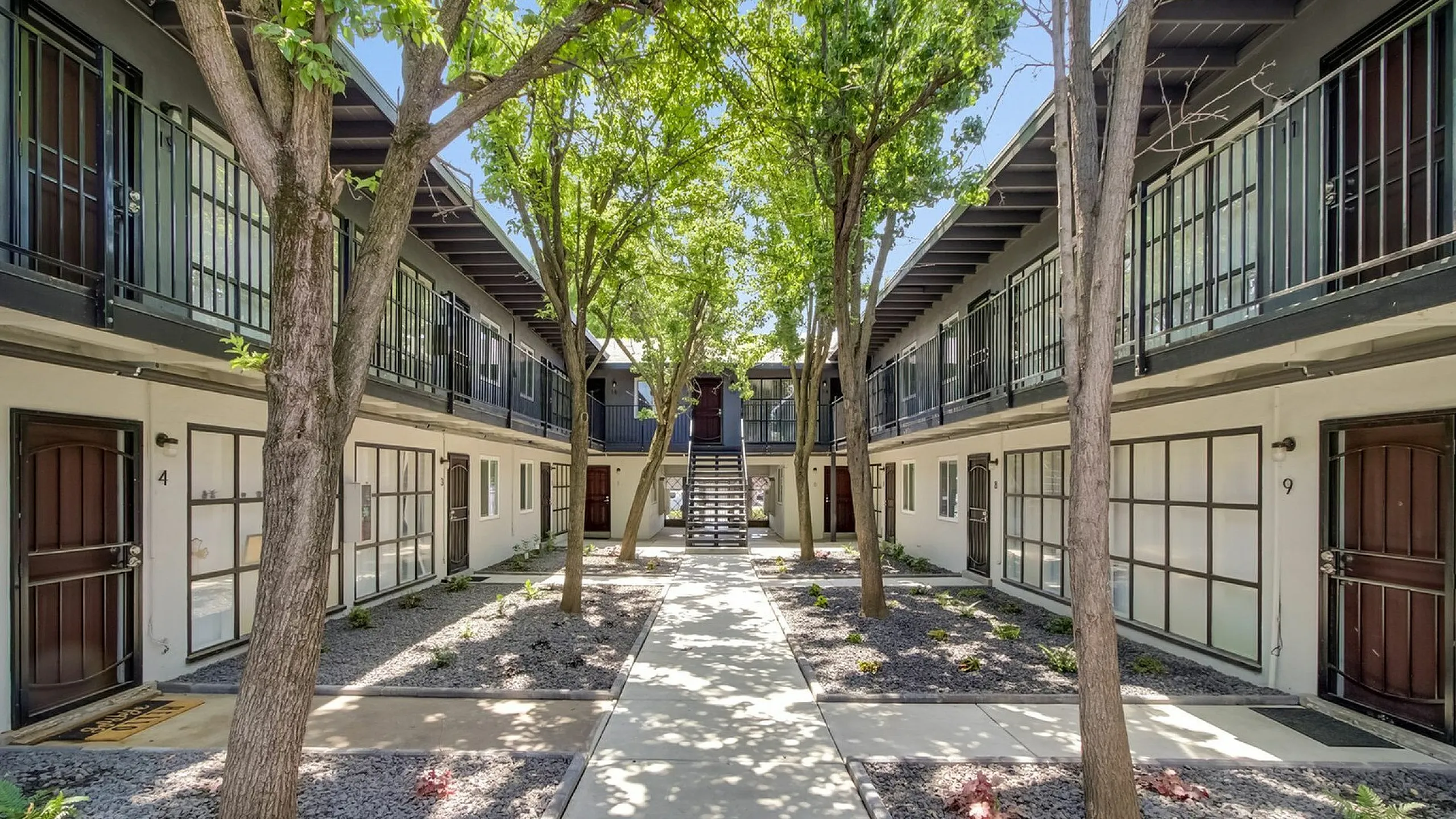 Two-story apartment complex with a central pathway lined by trees and gravel gardens. Sunlight filters through the leaves, creating a peaceful, inviting atmosphere.