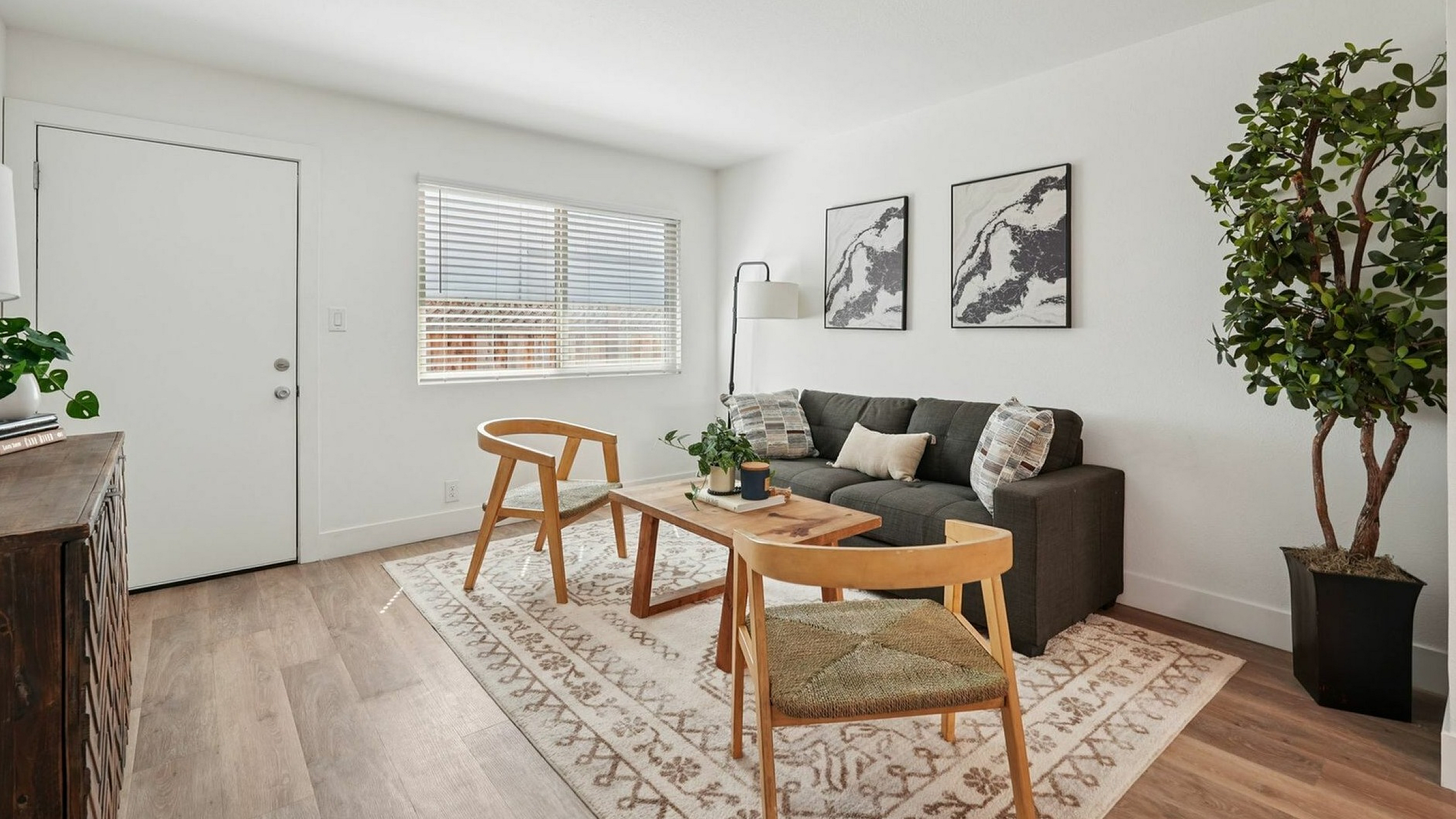 Modern living room with a cozy atmosphere. Features a gray sofa, wooden chairs, coffee table, large plant, and abstract art. Bright and inviting space.