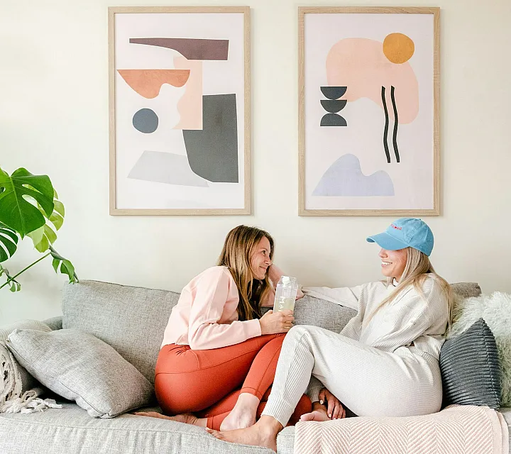 Two women sit on a gray sofa, smiling and chatting. Abstract art hangs on the wall behind them. A coffee table with books is in the foreground. Cozy and relaxed atmosphere.