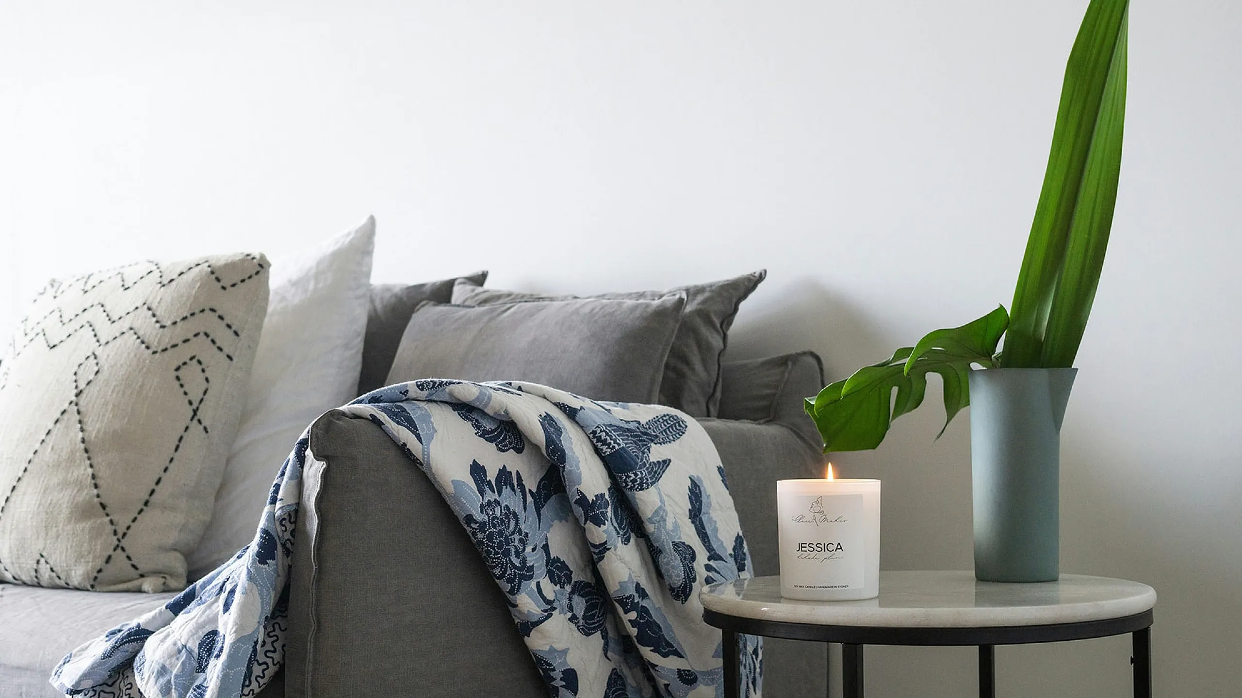 Cozy living room with a gray sofa, patterned throw and cushions. A lit candle and green plant in a vase rest on a round side table, creating a calm ambiance.