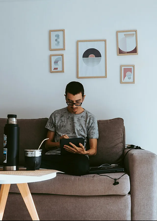 A person sits on a couch, focused on a tablet. Wall art and a bottle rest nearby. The scene is calm and cozy, with soft lighting and muted tones.