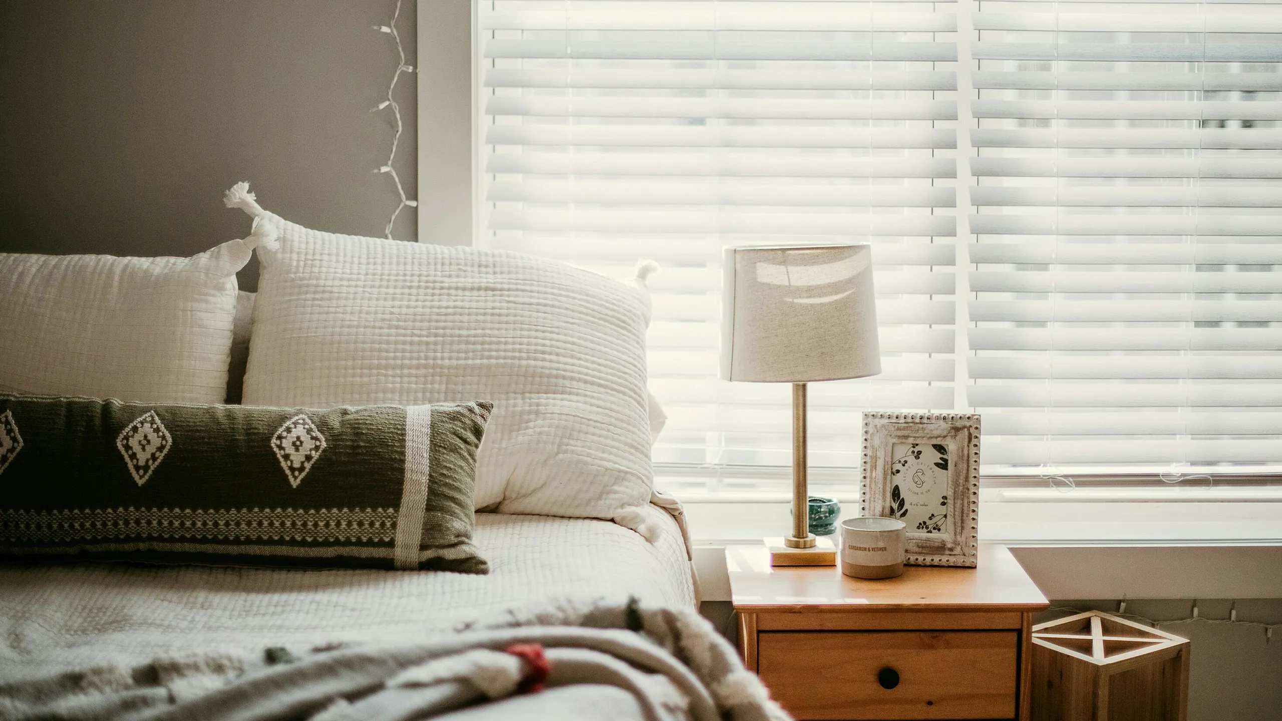Cozy bedroom with a neatly made bed, accented by white pillows and a patterned cushion. A wooden nightstand holds a lamp, framed photo, and small decor. Soft natural light streams through the blinds, evoking calmness.