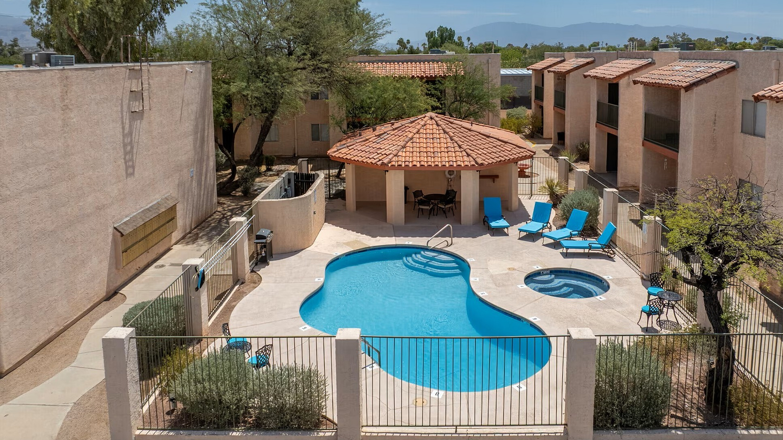 Sunny courtyard with a kidney-shaped pool and hot tub, surrounded by beige buildings and desert trees. Blue lounge chairs add a relaxing vibe.