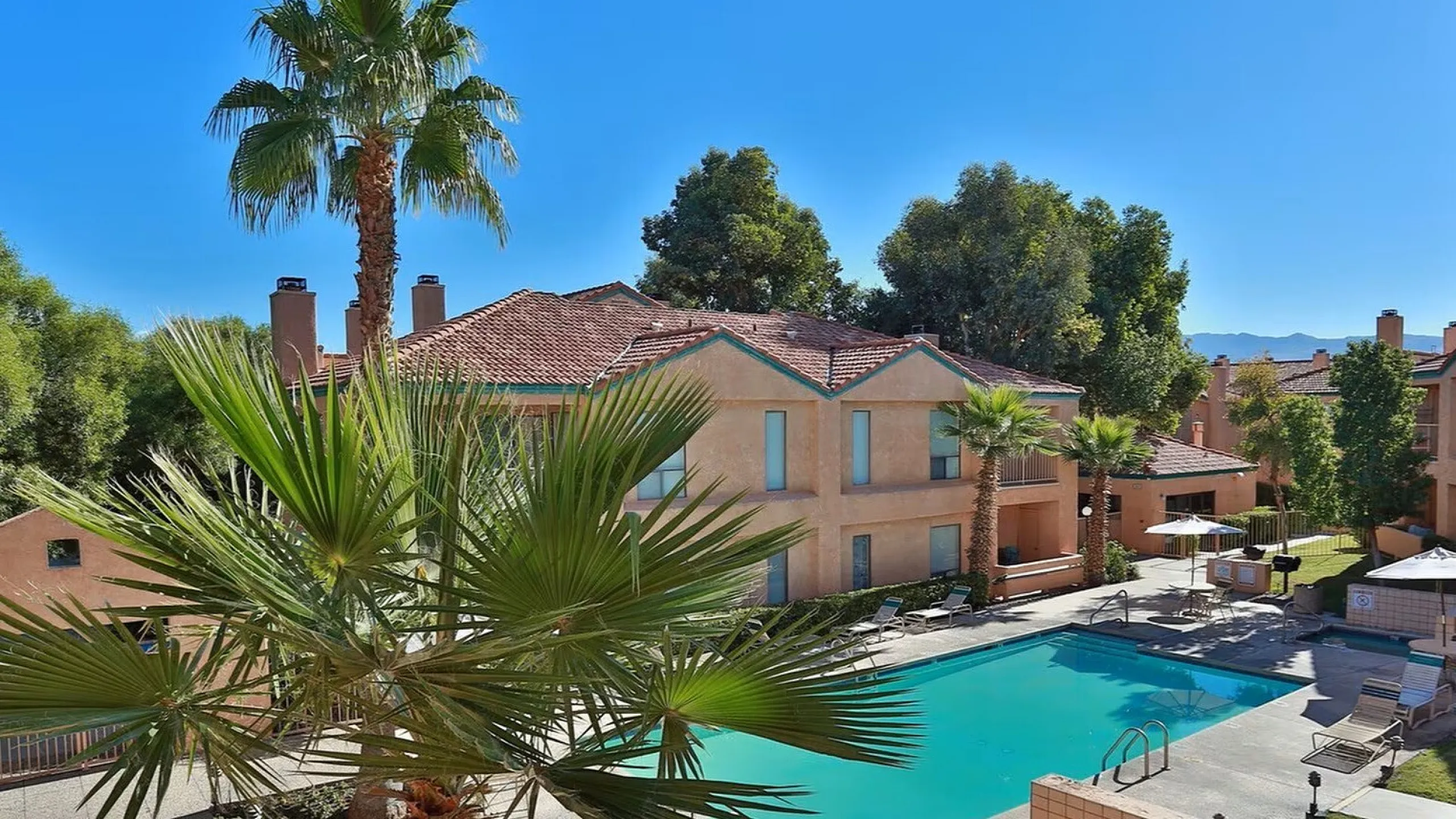 Sunny view of a serene outdoor pool surrounded by Mediterranean-style buildings with red-tile roofs. Palm trees and lounge chairs create a relaxed atmosphere.