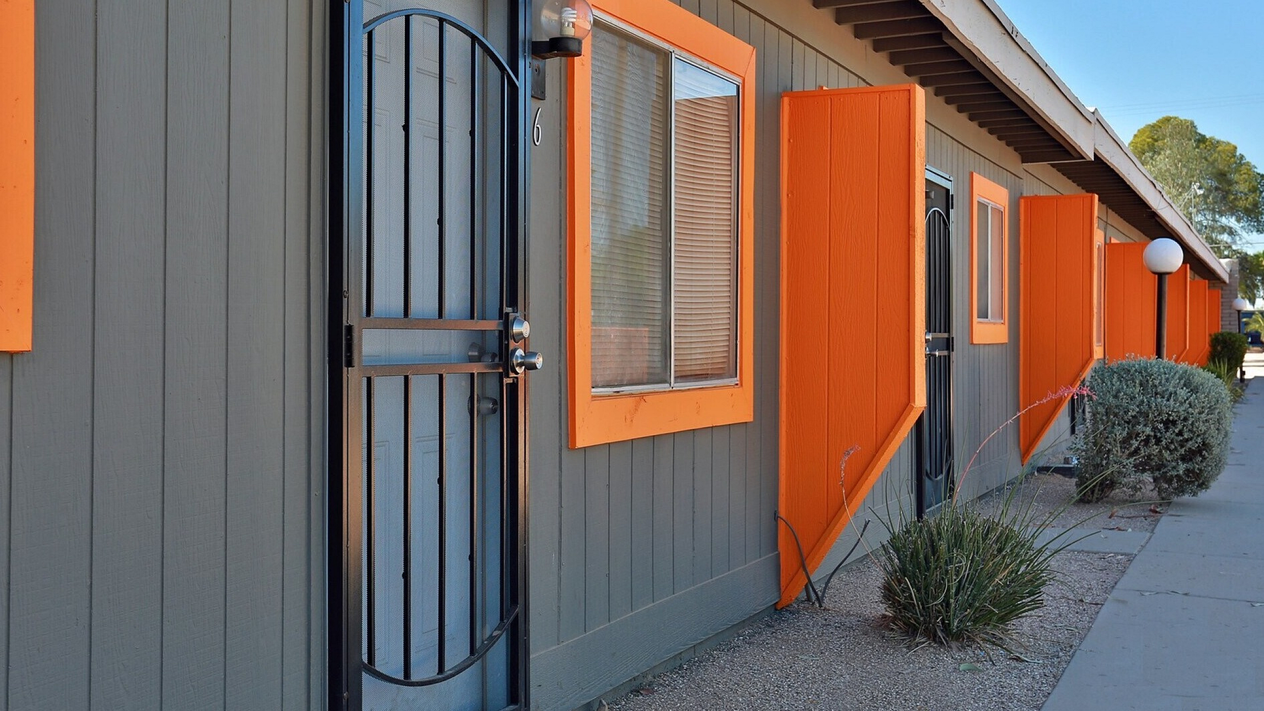 A row of small gray buildings with bright orange window frames and shutters. A black metal door is visible on the left. The scene is sunny and neat.