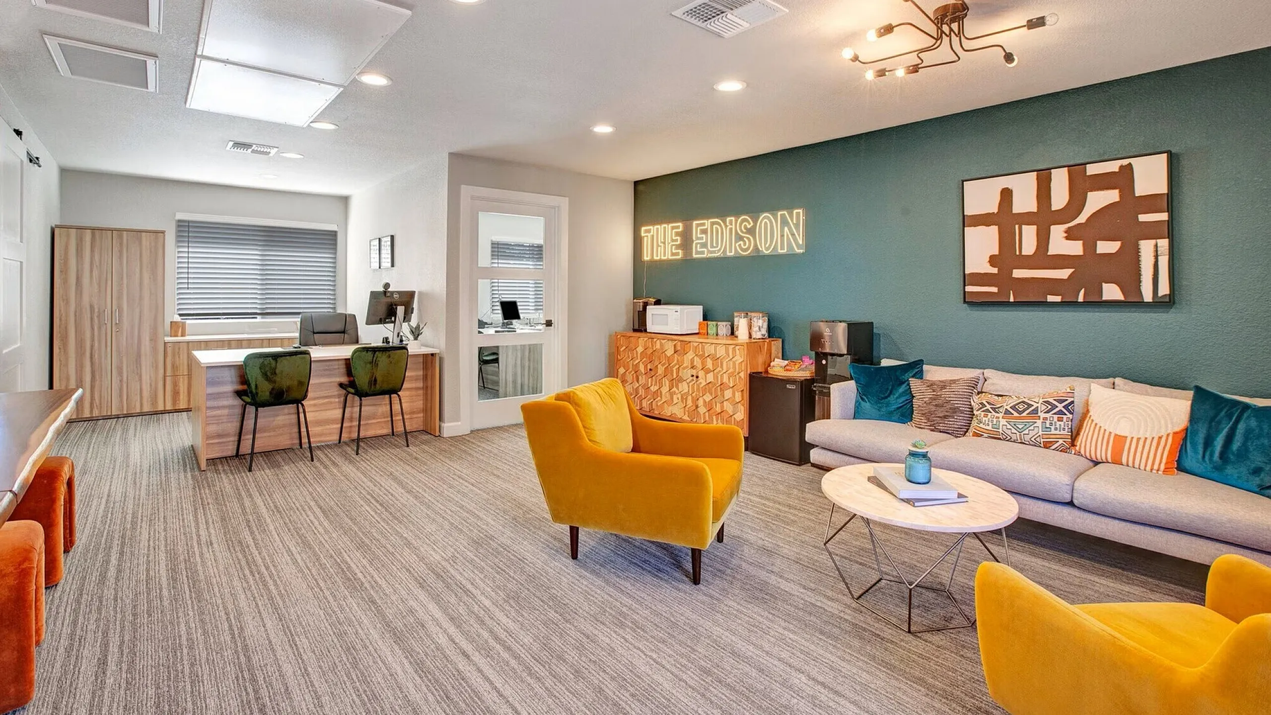 Modern office lounge with vibrant decor. Yellow chairs, gray sofa, teal accent wall, abstract art, and neon sign "The Edison." Cozy, inviting atmosphere.