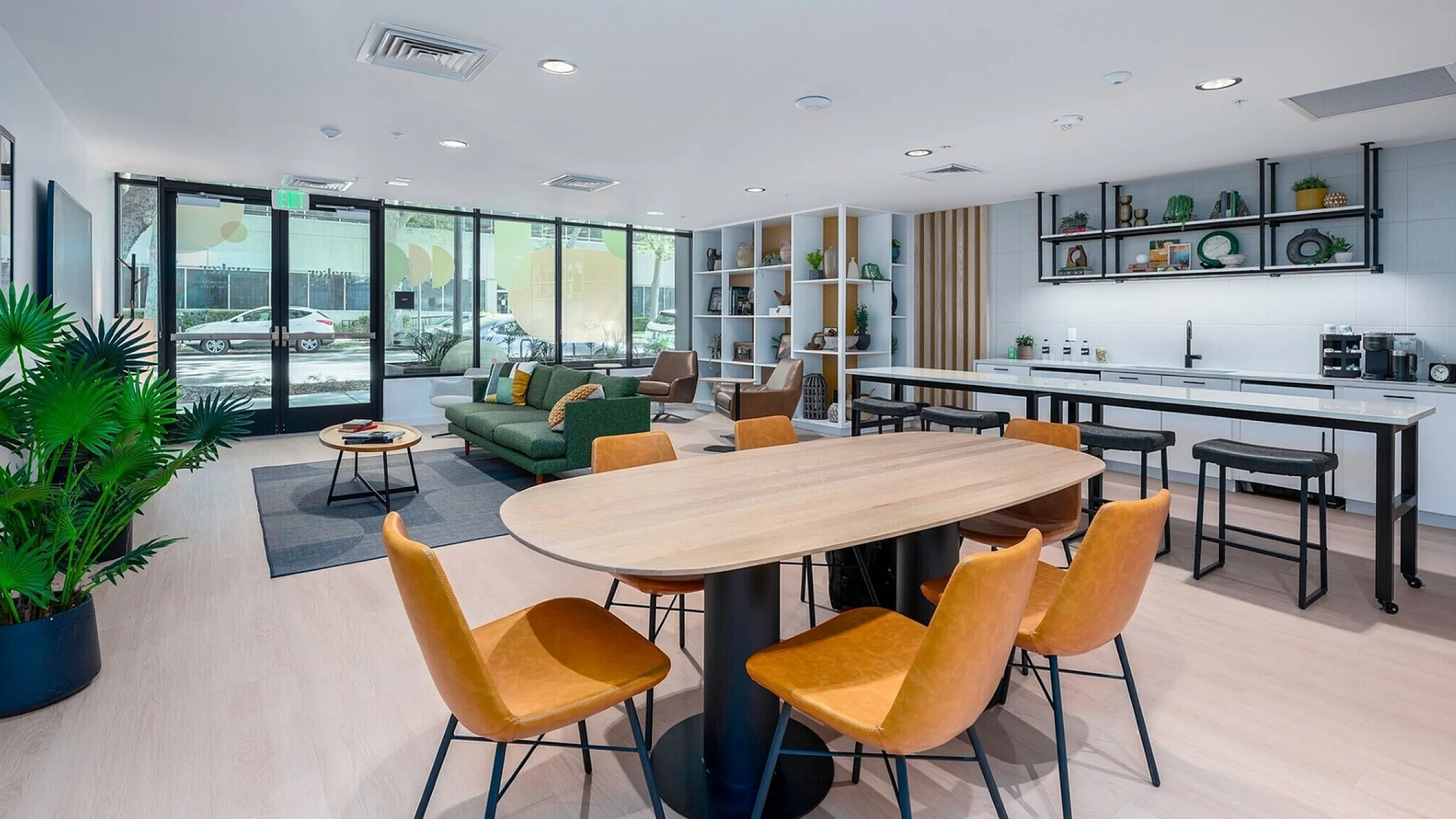 Modern lounge area with a large oval table and six orange chairs. Nearby, a green sofa and armchairs sit on a gray rug. The room is bright, with plants and a sleek kitchen.
