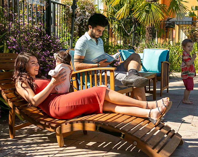 Man reads on a lounge chair by a pool, while a woman smiles, holding a baby. The scene is sunny and relaxed.