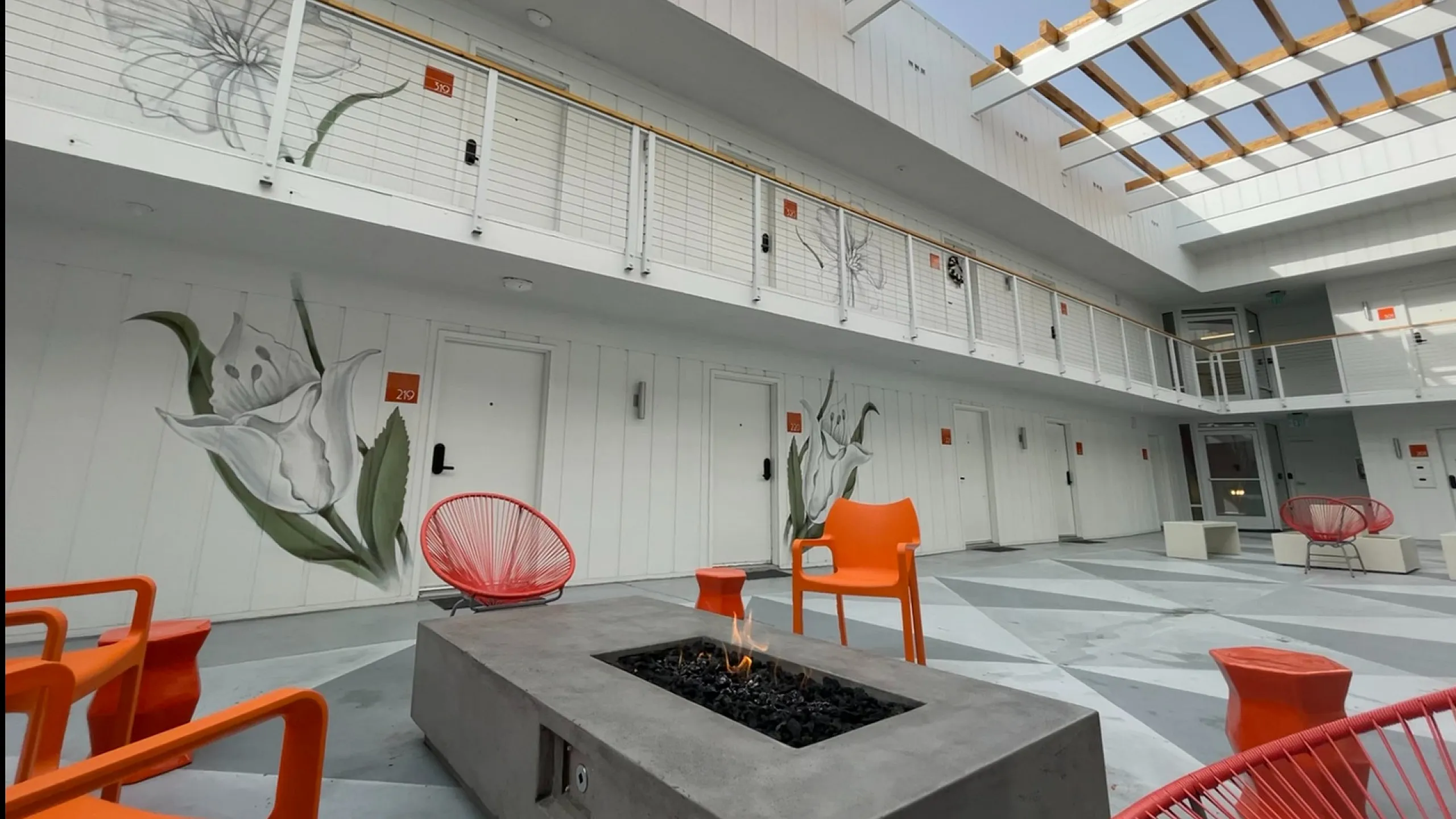 Modern courtyard with vibrant orange chairs surrounding a central fire pit. White walls feature large floral murals. The open roof design adds a bright, airy feel.
